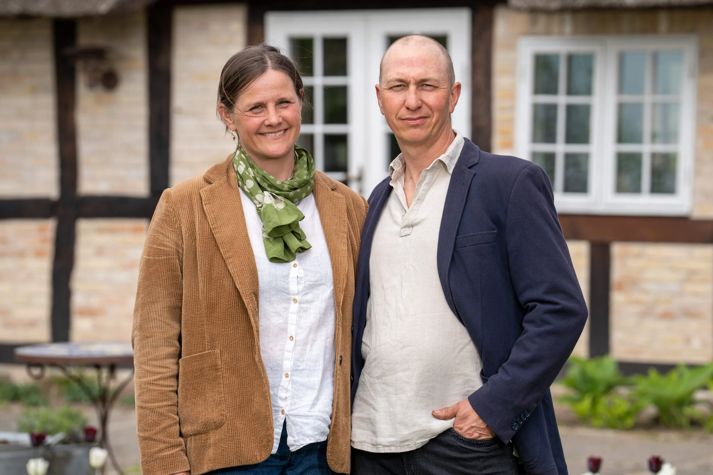 A woman with brown hair wearing a green scarf, white shirt, and brown blazer standing next to a man with short hair wearing a light-colored shirt and navy blazer, outdoors in front of a house with large windows.