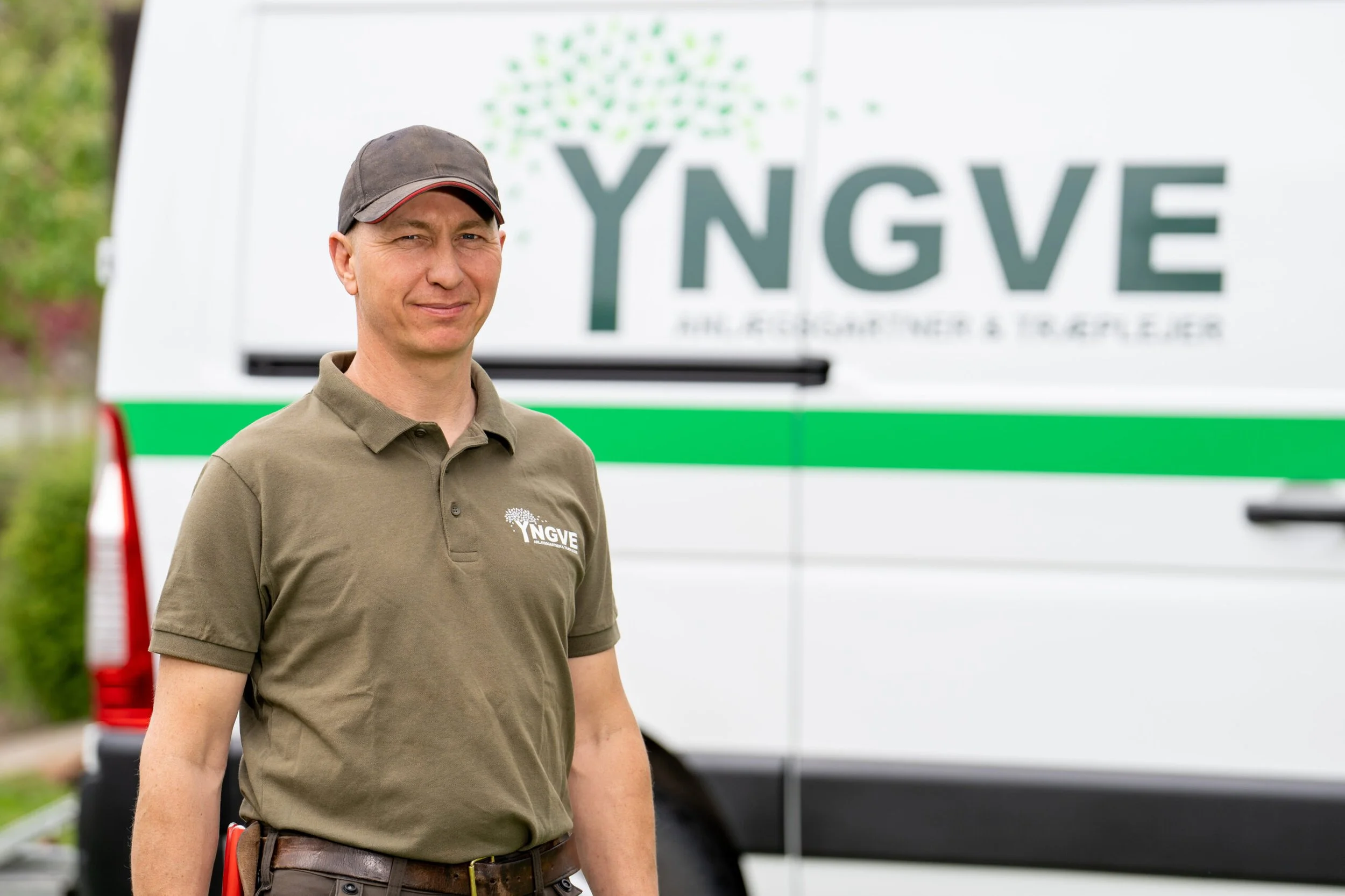 En mand iført en grøn polo med NGVE logo står foran en servicebil med NGVE logo i baggrunden.