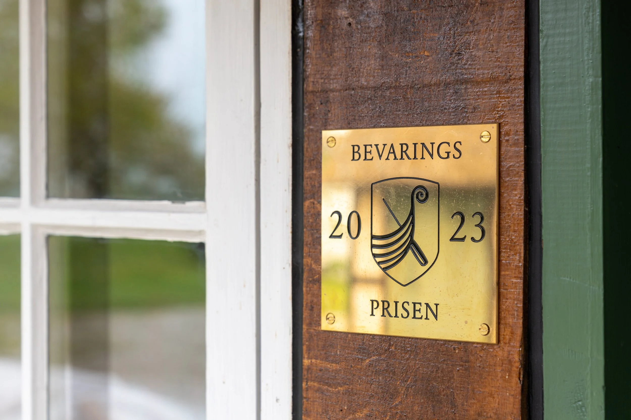 Close-up of a brass sign on wooden exterior wall with the word 'BEVARINGS' and the numbers '20' and '23'.