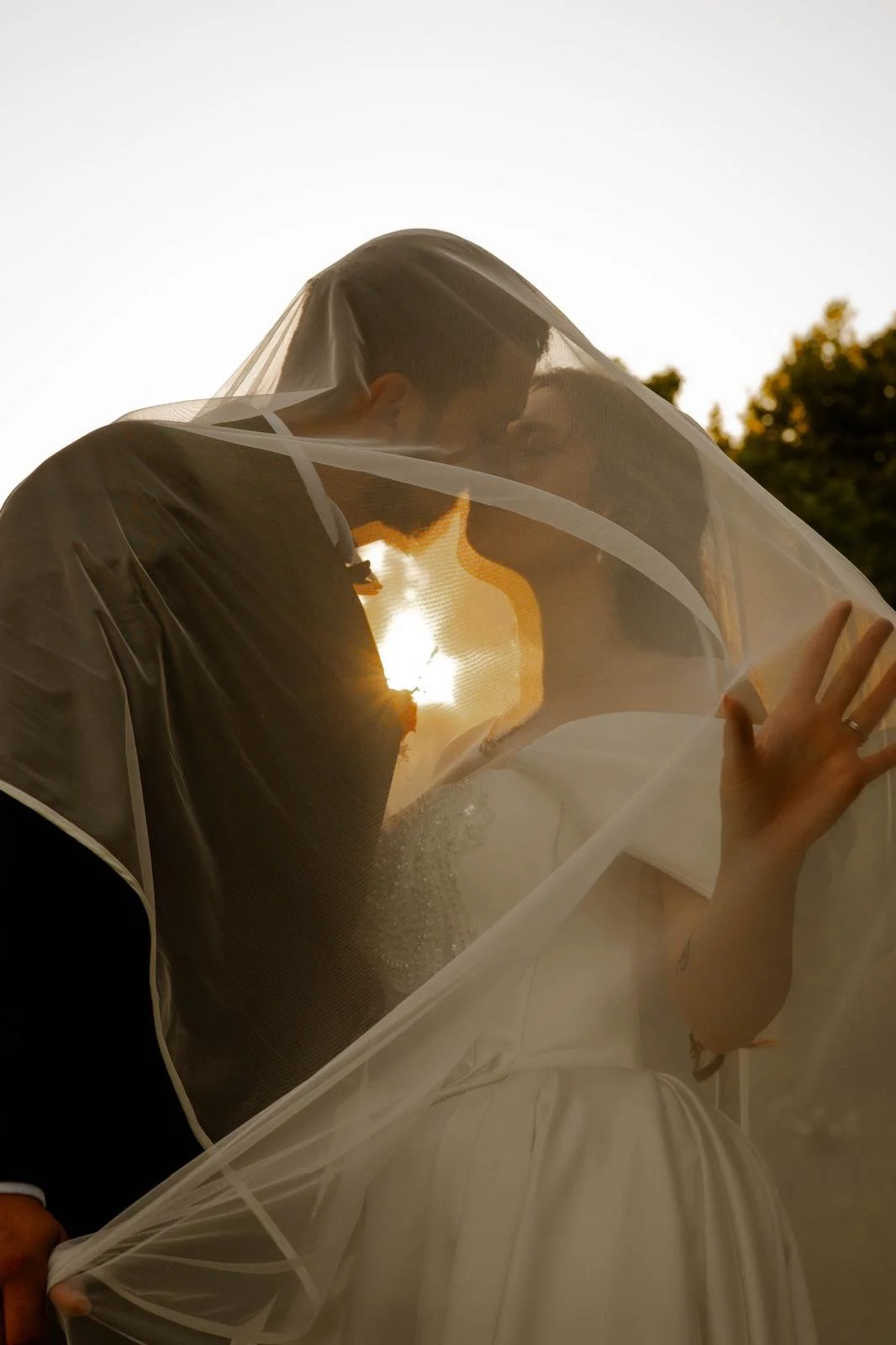 A couple dressed in wedding attire sharing a kiss under a veil, with the sun setting behind them and trees in the background.
