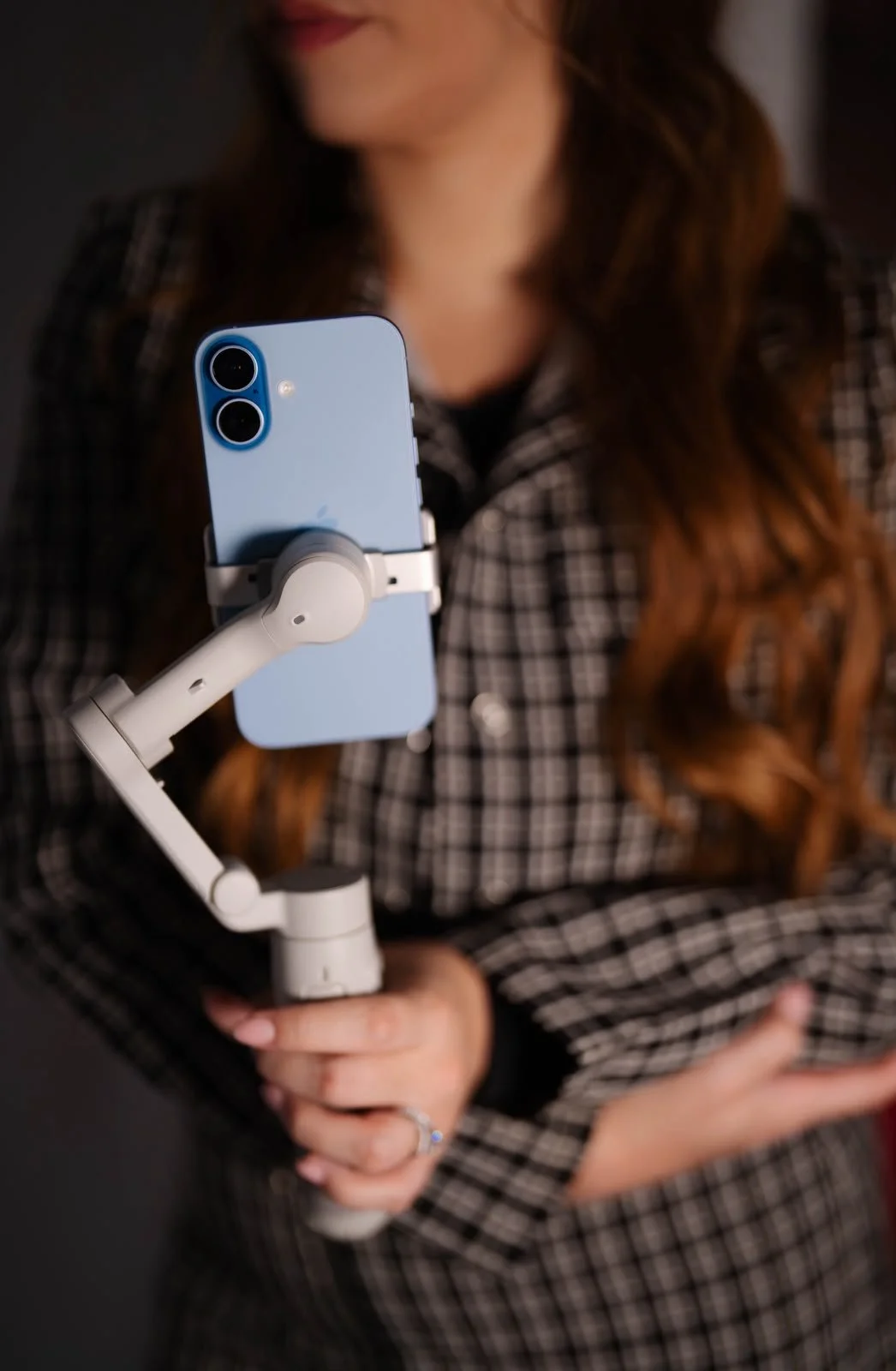 A woman with long, wavy red hair wearing a black and white plaid shirt is taking a selfie with a light blue iPhone 13 using a white handheld stabilizer or selfie stick.