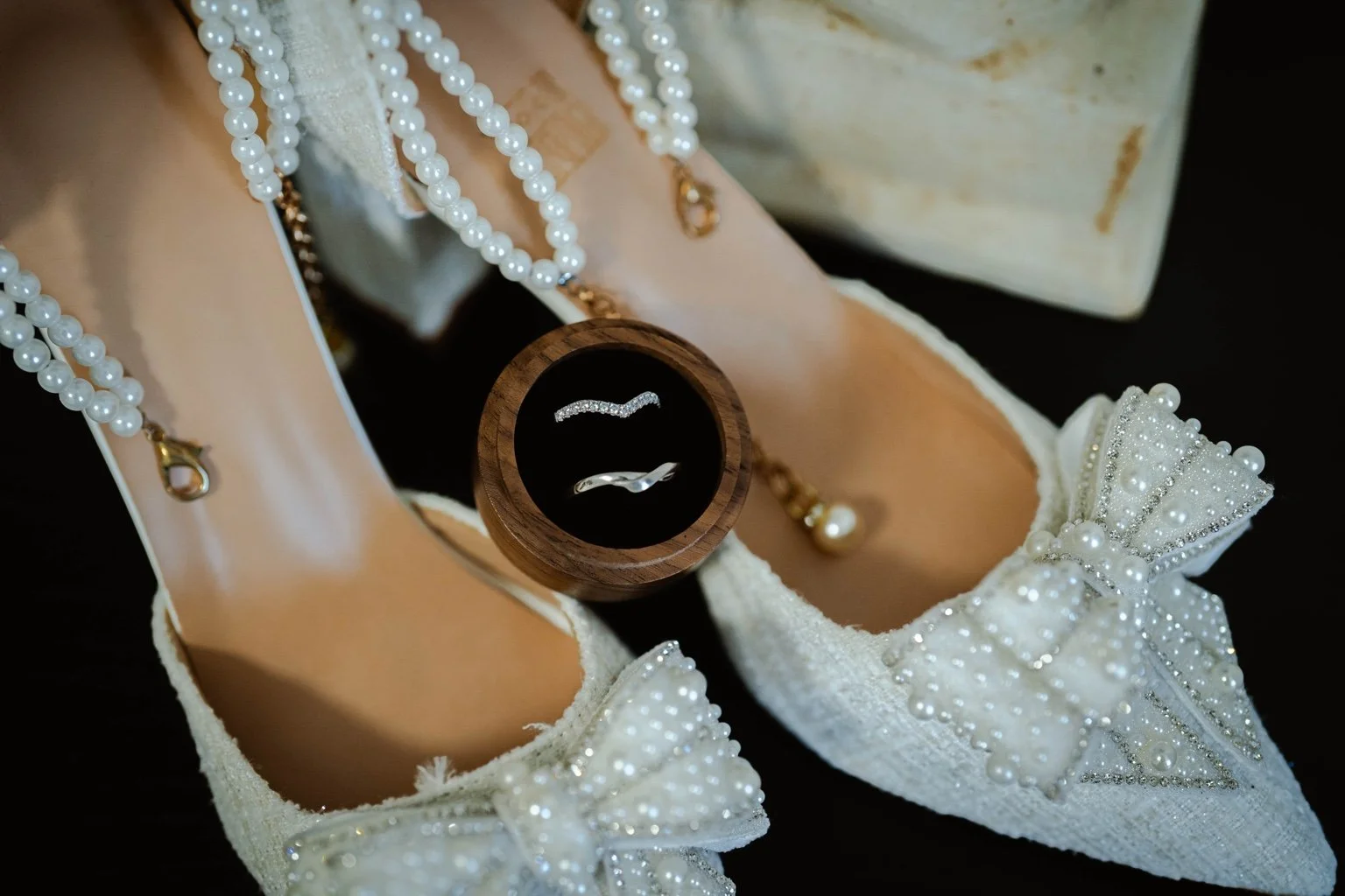 A pair of wedding shoes decorated with pearls and bows, with a pearl necklace, rings, and jewelry inside a wooden ring box.