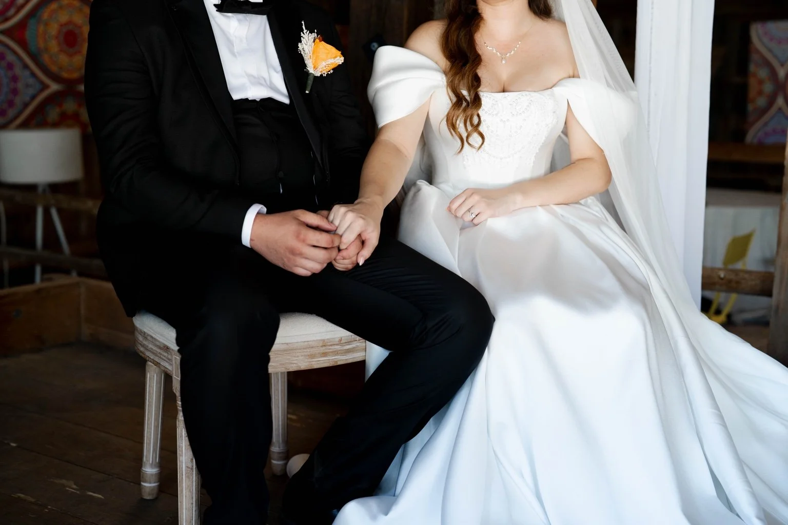 A bride and groom sitting together, holding hands, during their wedding ceremony. The bride is wearing a white wedding gown with off-the-shoulder sleeves and a veil, and the groom is dressed in a black suit with a white shirt and a boutonniere.