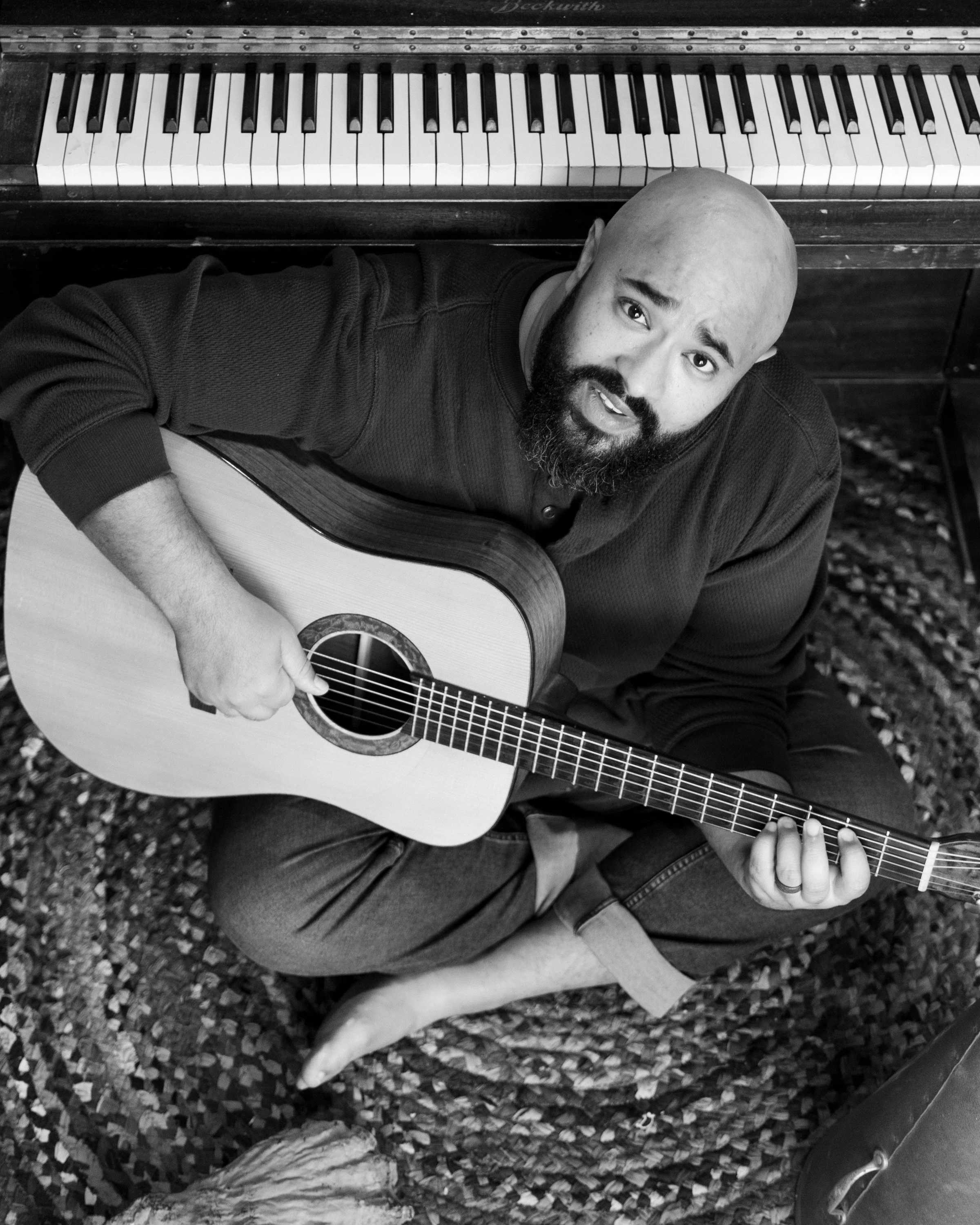 A man looking up while seated on the floor playing a guitar in front of a piano