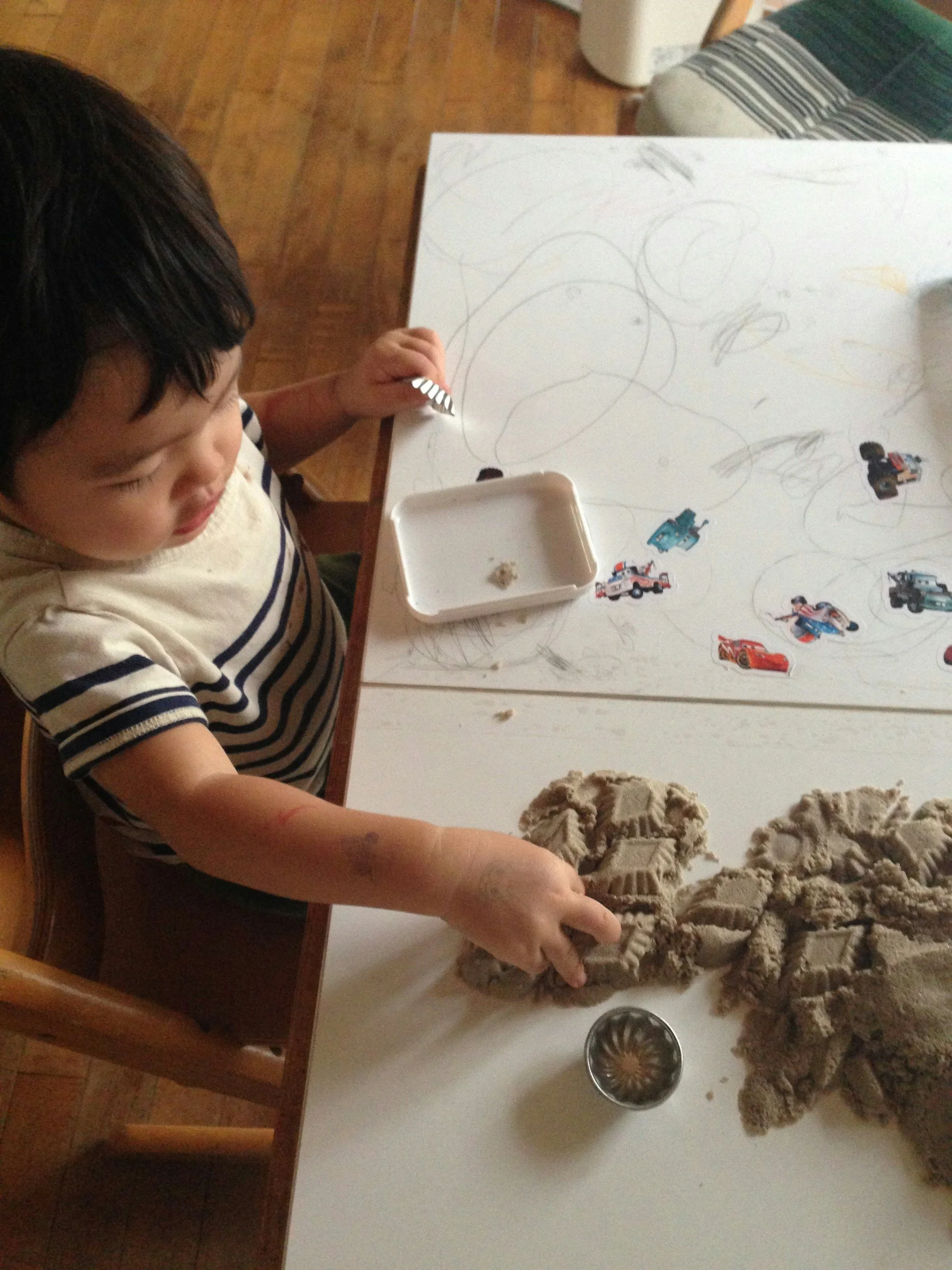 A young boy with dark hair, wearing a striped shirt, is playing with beige modeling clay on a white table. The table has sketches and stickers of cars on it, and there are some scattered clay pieces and a metal container nearby.