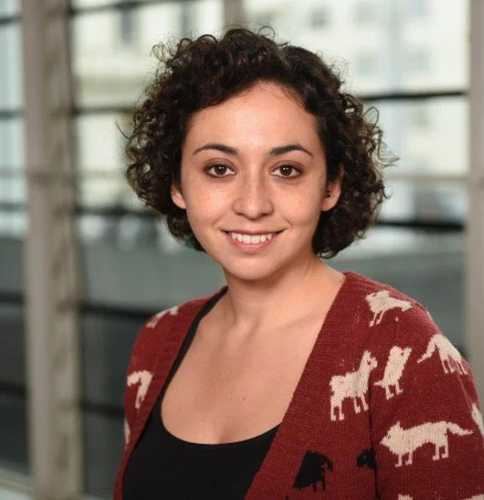 Maria Crespo-Ribadeneyra, a young woman with short, curly hair smiling at the camera. She is wearing a black top and a red cardigan with white animal patterns, standing indoors. Maria is a battery researcher at Queen Mary's University.