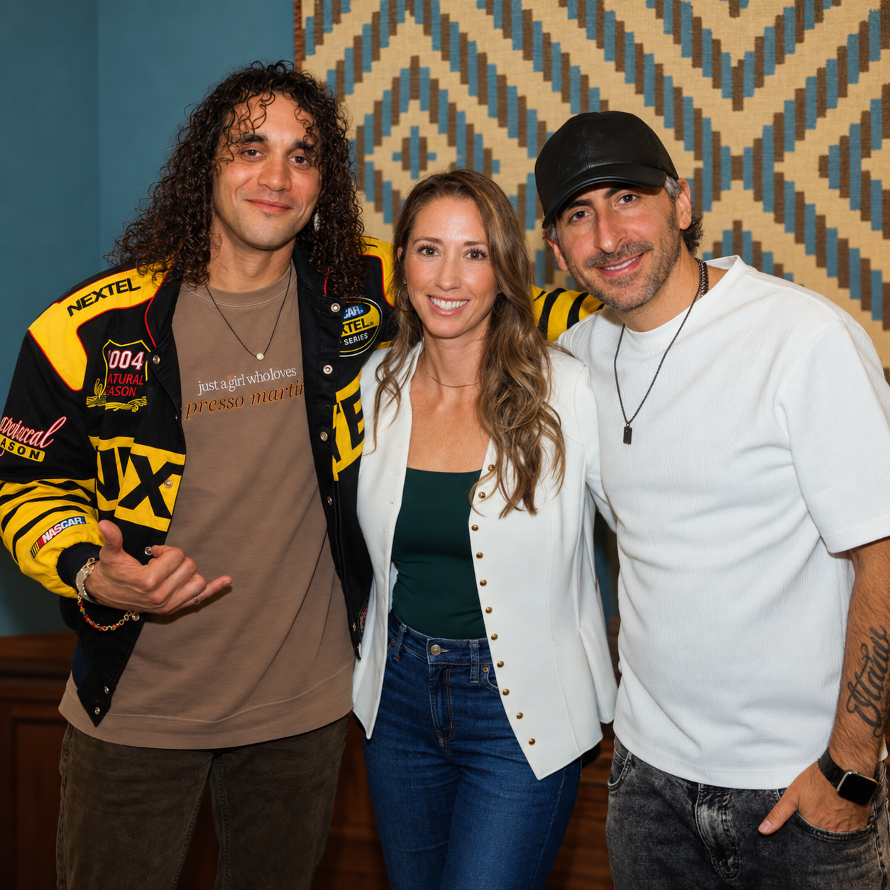 Three people standing close together in a room, smiling at the camera. The person on the left has curly hair, wearing a racing jacket and a t-shirt that says 'just a girl who loves presso martini.' The woman in the middle has long wavy hair, wearing a white blazer over a green top. The person on the right has a tattoo on his forearm, wearing a white shirt, a black cap, and a necklace.