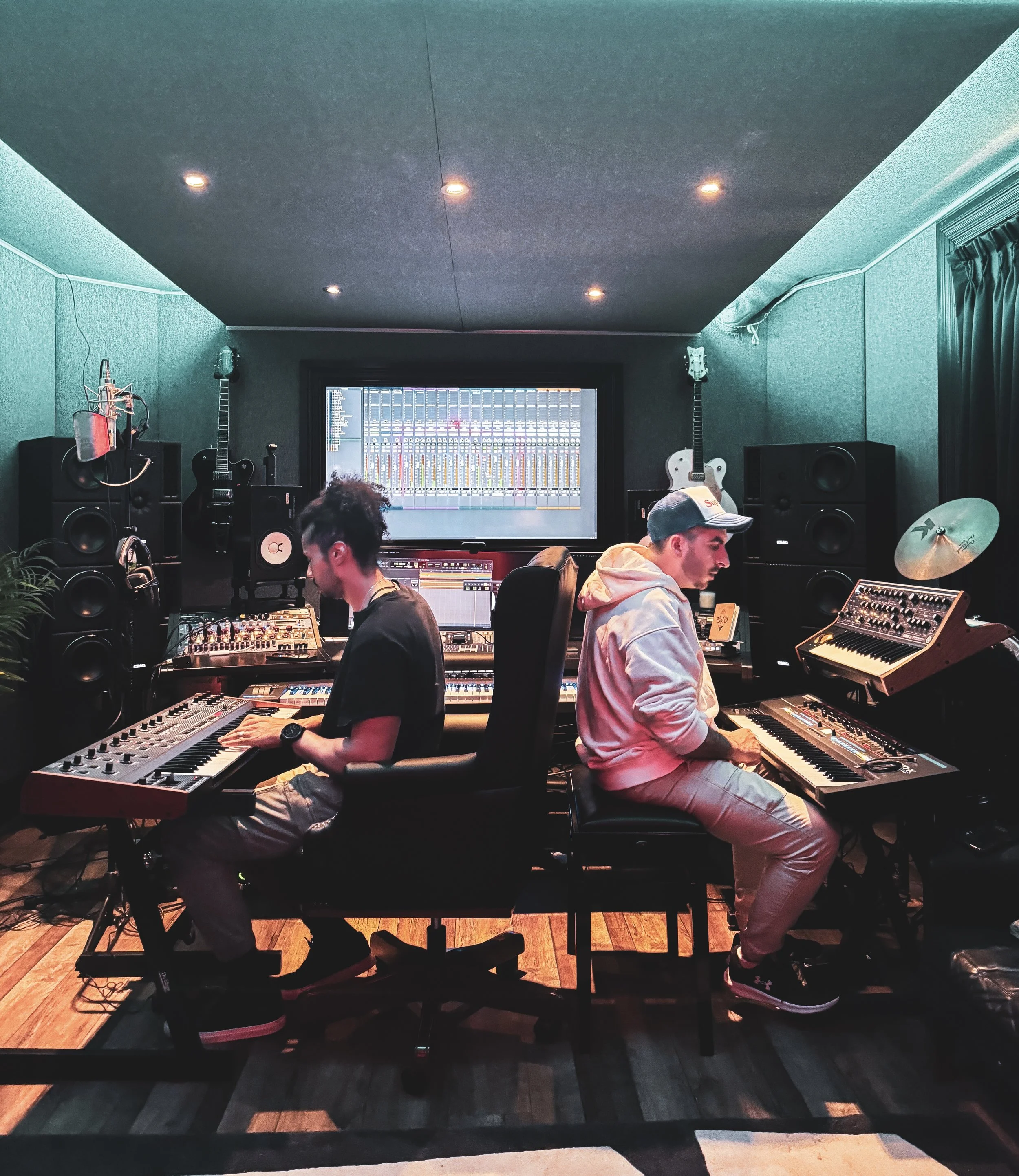 A music studio with two people working at a mixing console, surrounded by speakers, guitars, and music equipment. A large screen displays audio mixing levels.