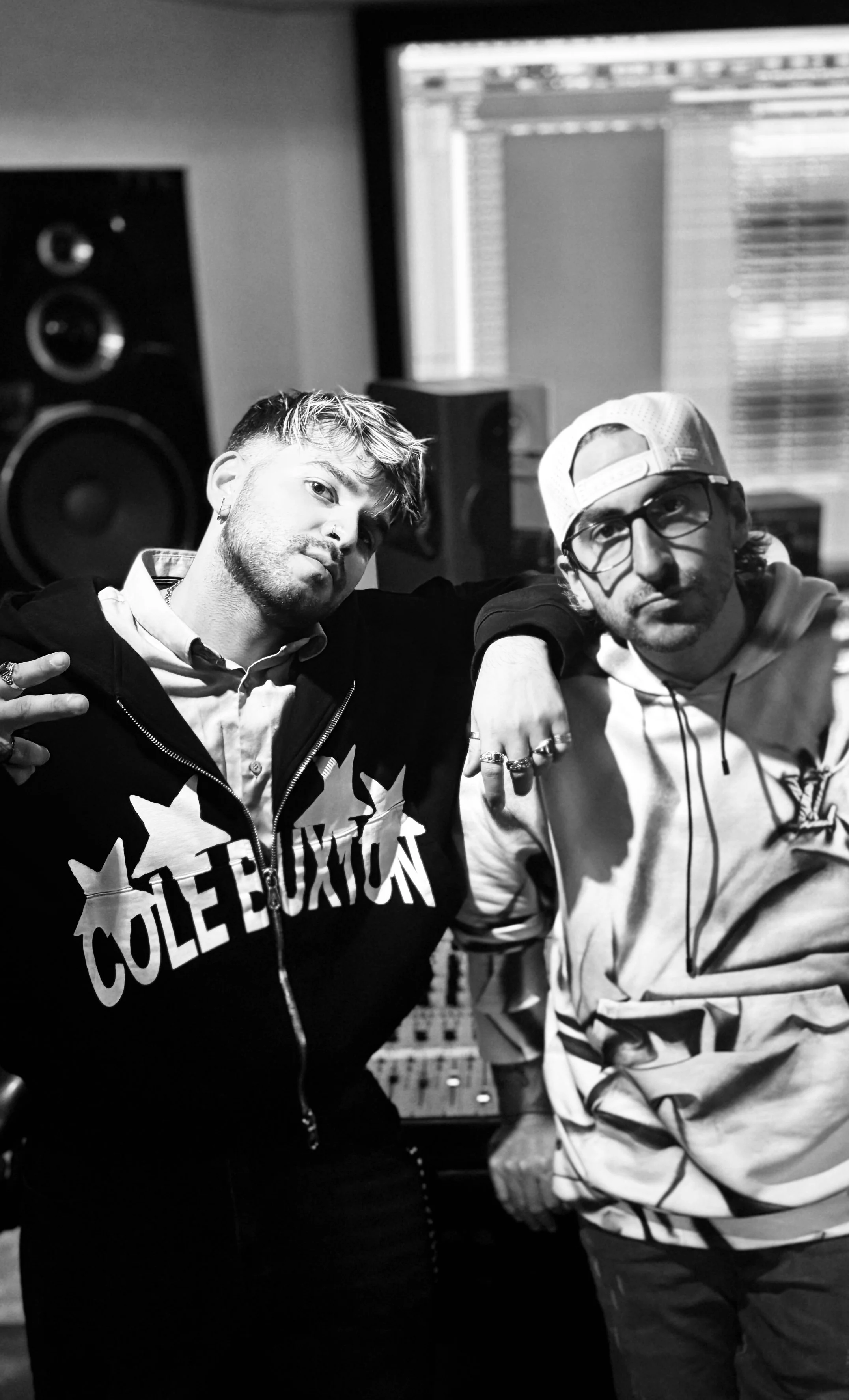 Two young men sitting closely together in a music studio with audio equipment, one wearing a hoodie with large text and the other wearing glasses, a cap, and patterned jacket.