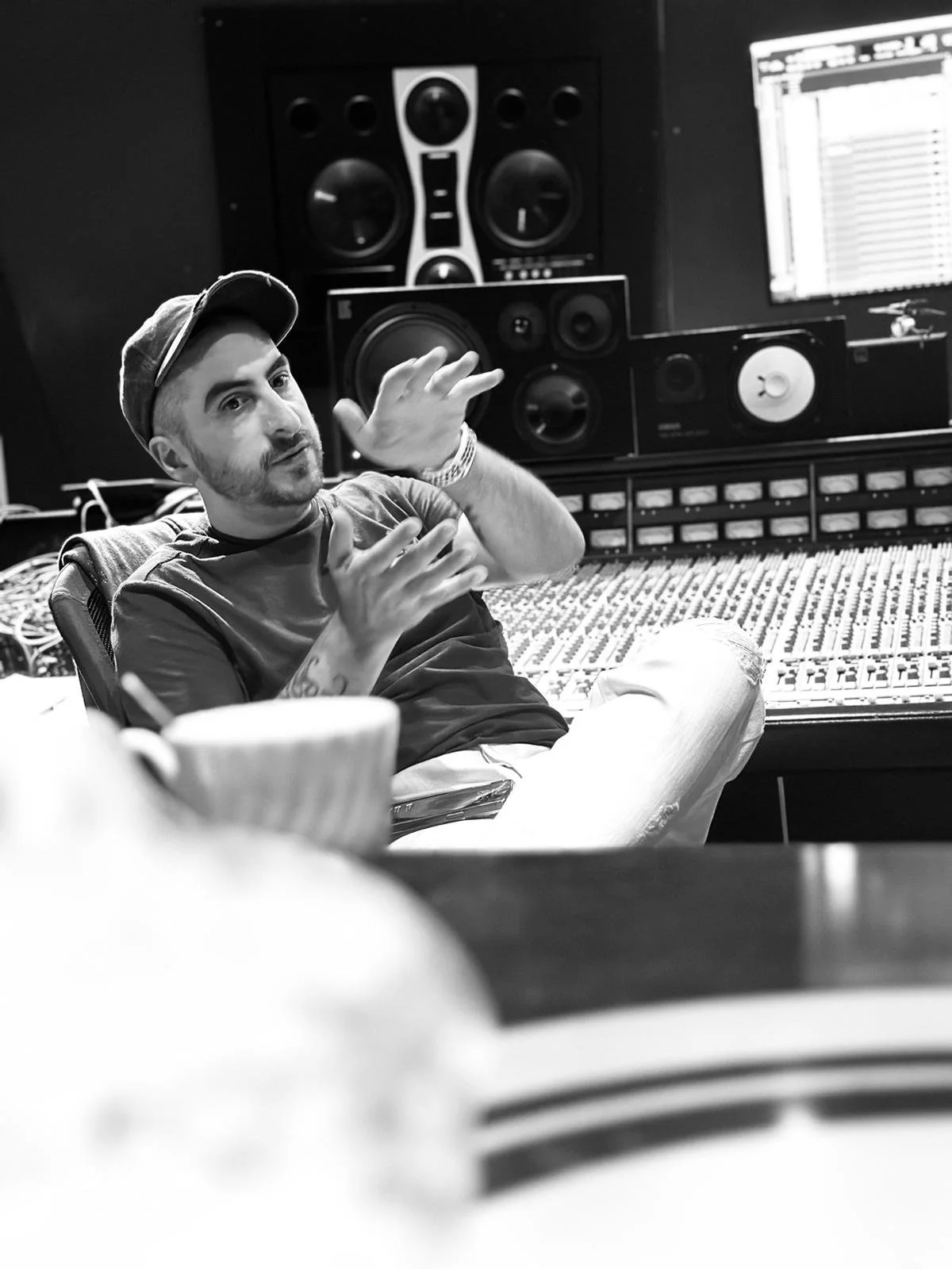 A man wearing a cap gestures with his hands while sitting in a recording studio with professional audio equipment and sound mixing console.