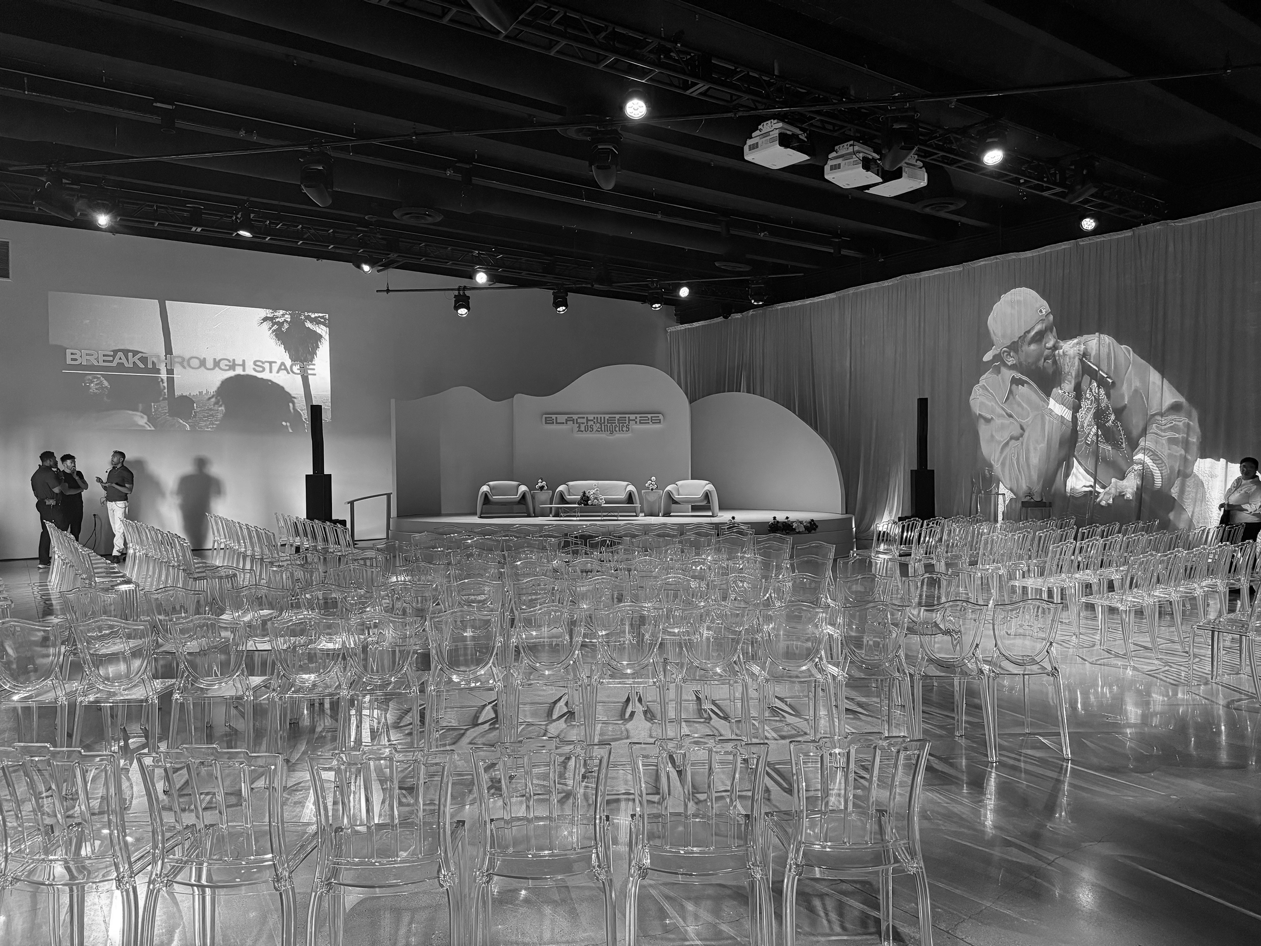 An empty stage set up for a panel or presentation with three chairs and small tables, a large screen in the background displaying a palm tree and the text 'BREAKTHROUGH STAGE,' and a puppet on the right side of the stage.