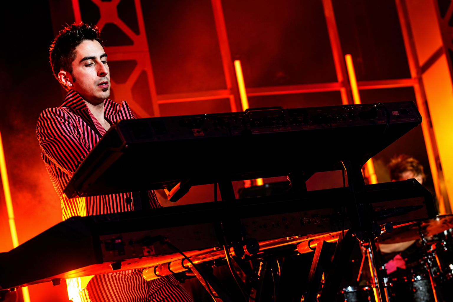 A musician playing keyboard on stage with red lighting and geometric background.