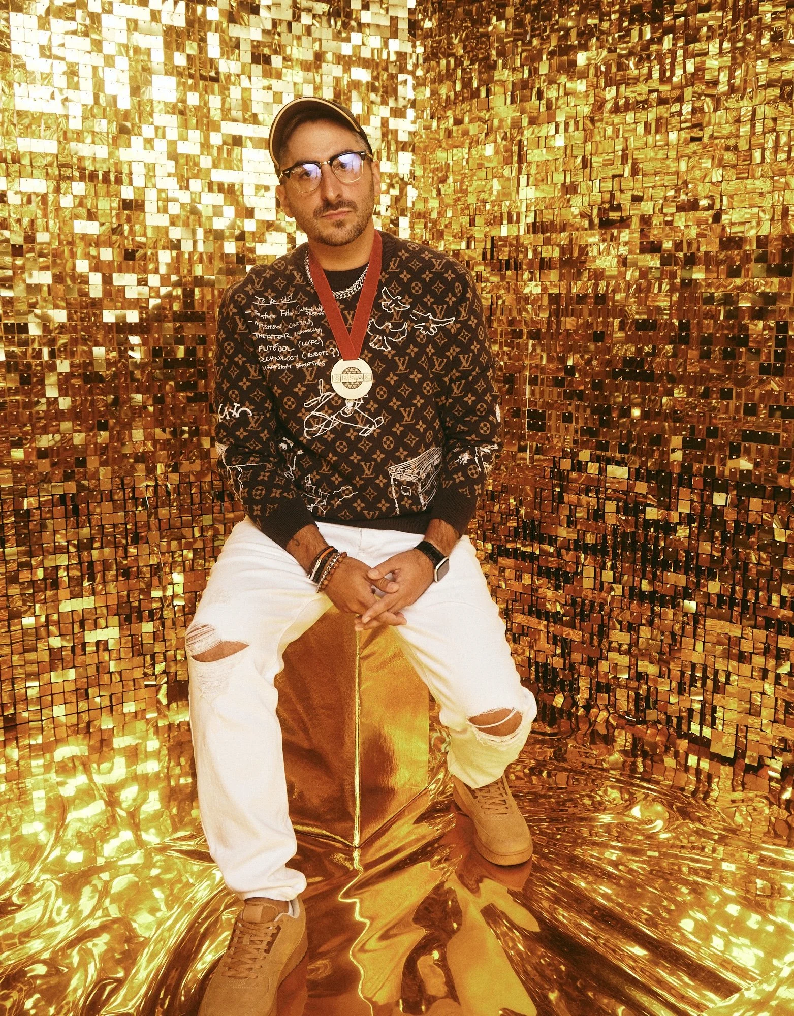 A man wearing glasses, a baseball cap, a patterned shirt, and white ripped jeans, sitting on a golden, reflective chair in front of a sparkling golden mosaic wall.