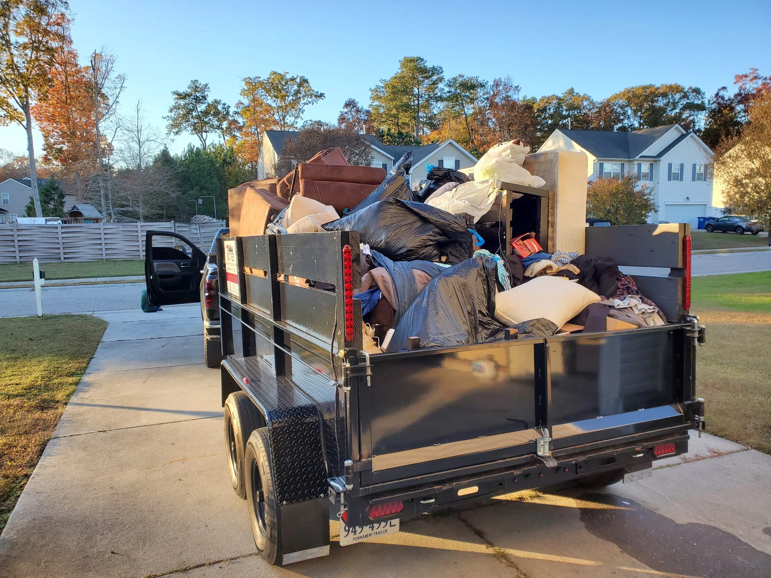 A trailer filled with various household items, including furniture and bags, parked on a driveway in a suburban neighborhood during daylight hours.