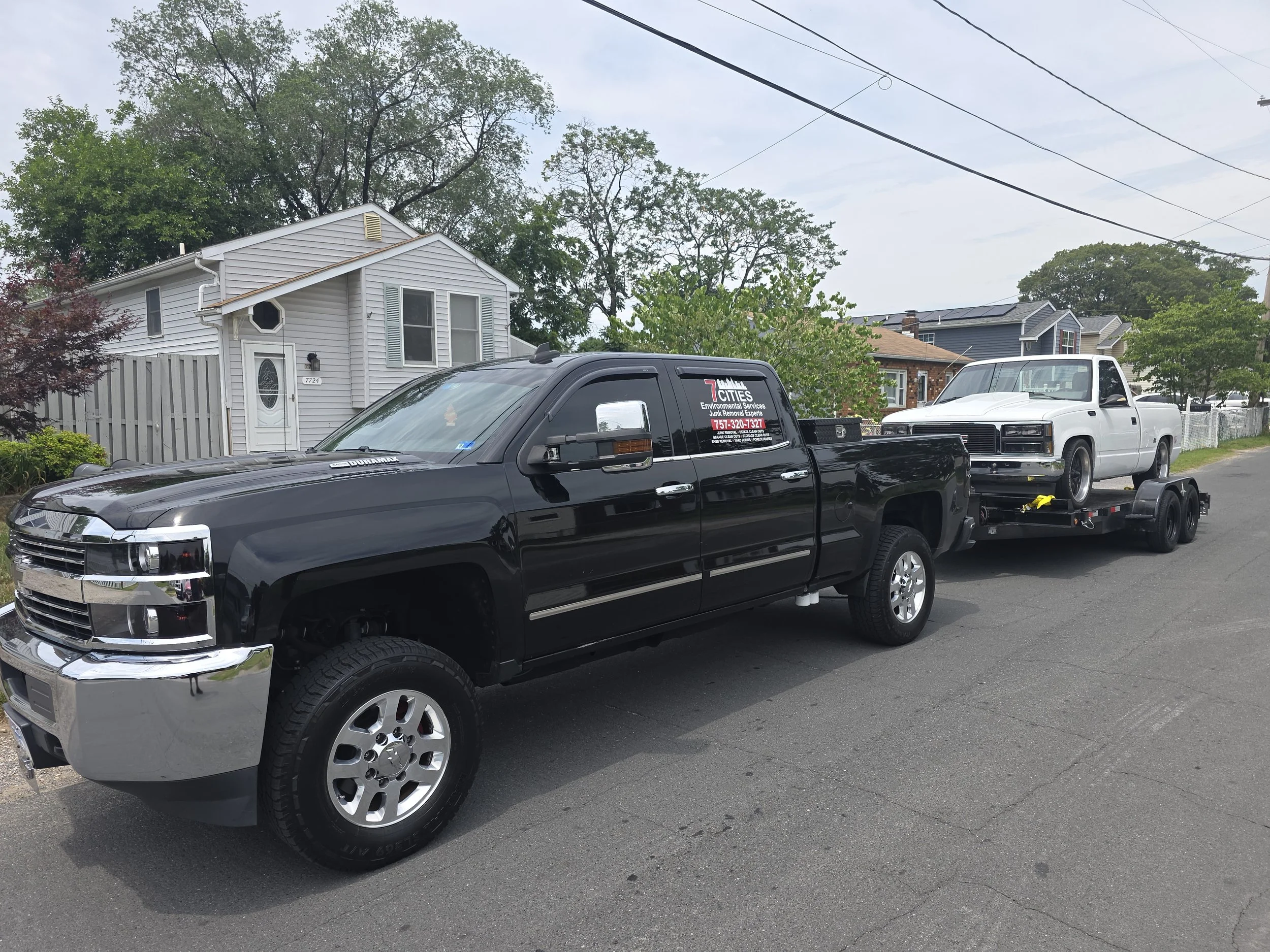 A black pickup truck on a street with a white house and another vehicle on a trailer parked nearby. Trees and power lines are visible in the background under a cloudy sky.