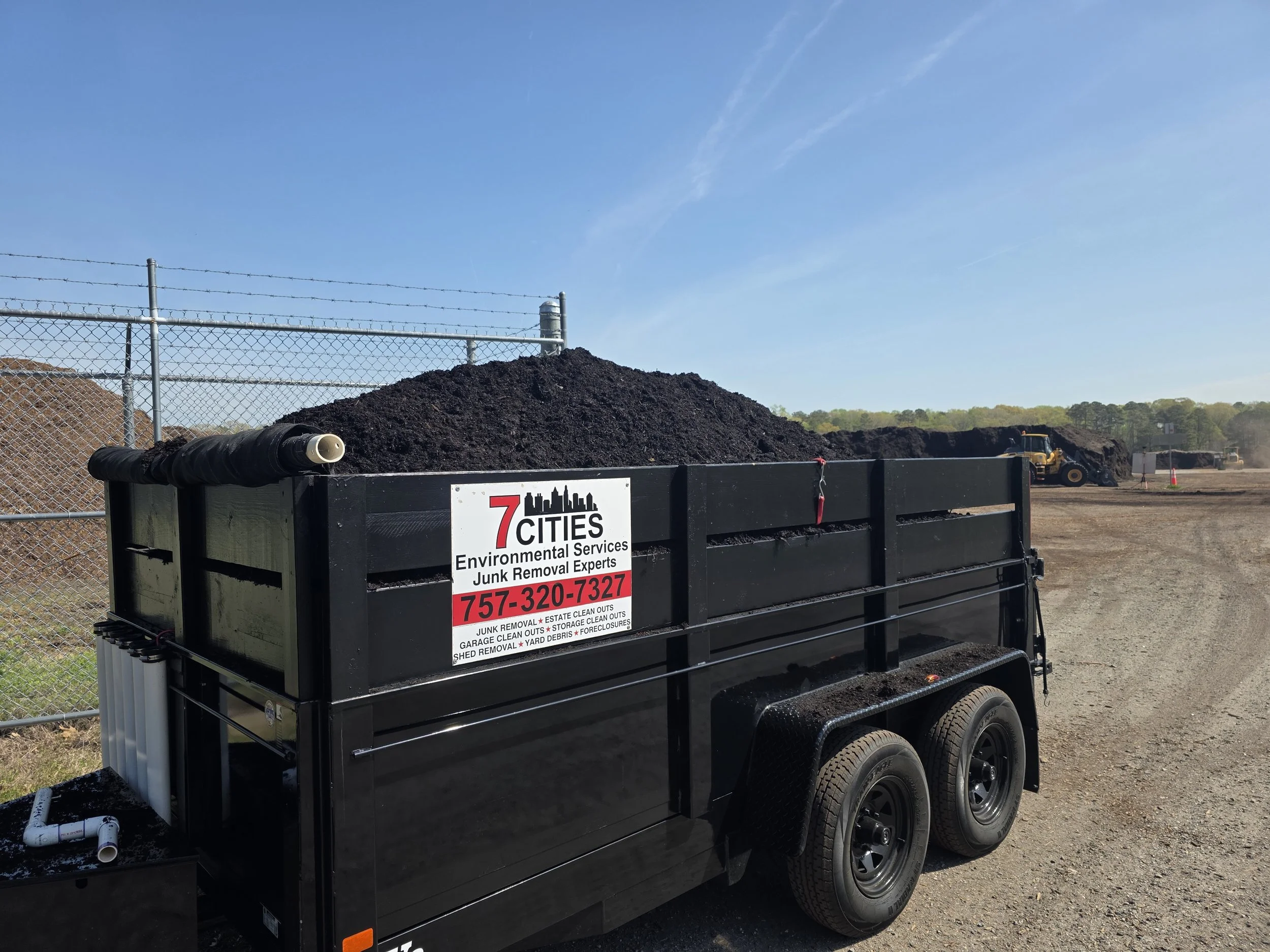 A black trailer filled with dark soil parked at a construction site, with a yellow bulldozer in the background, a metal chain-link fence, and a clear blue sky overhead.