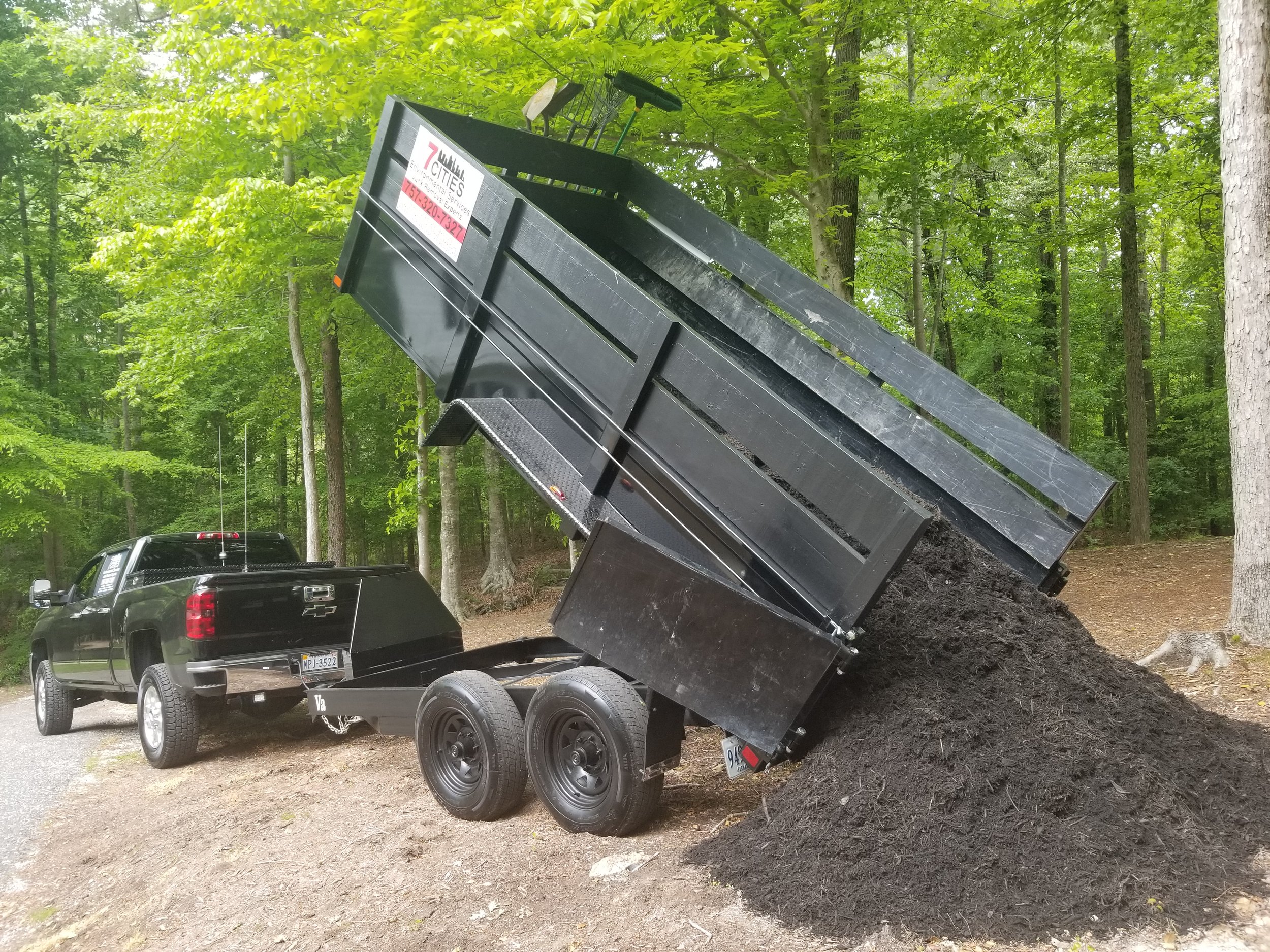 A black dump trailer attached to a black pickup truck is unloading dirt into a woodland area with green trees and soil.