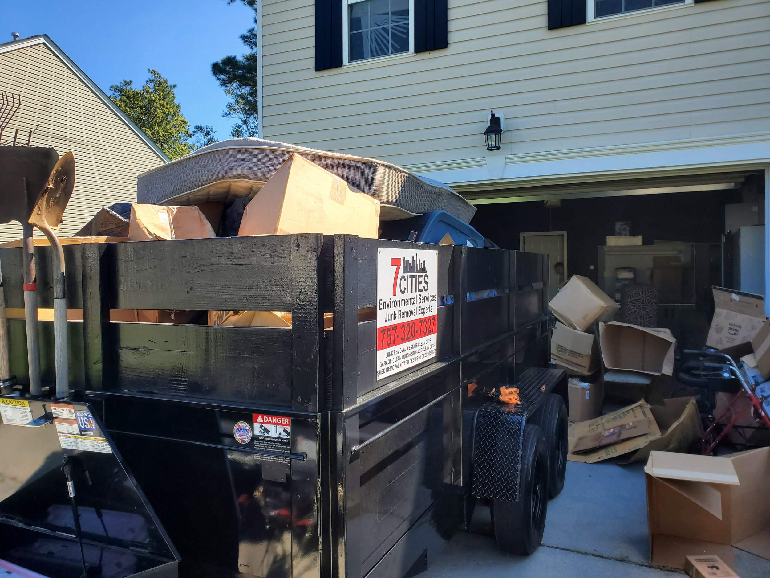 A black trailer filled with various items like a mattress, boxes, and cardboard, parked in a driveway beside a beige house with black shutters and a black outdoor wall lamp.