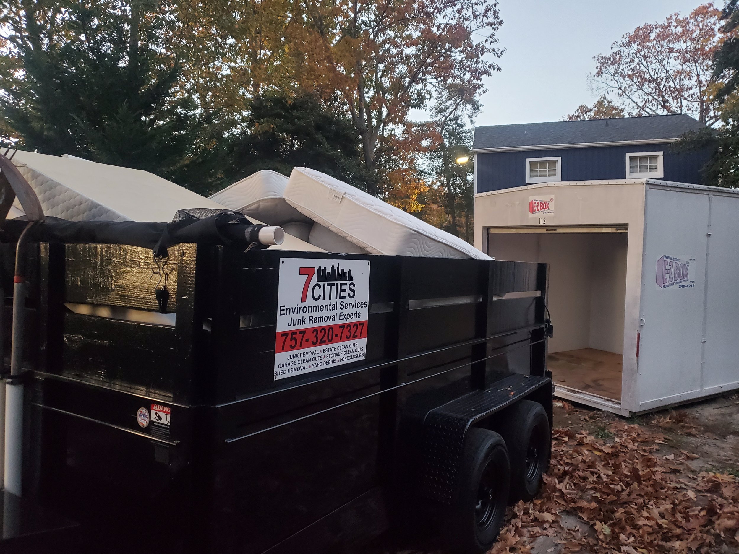 A black dumpster filled with mattresses and other junk items, parked in front of a blue house with a garage, surrounded by fallen leaves. There is a sign on the dumpster for environmental services and junk removal.