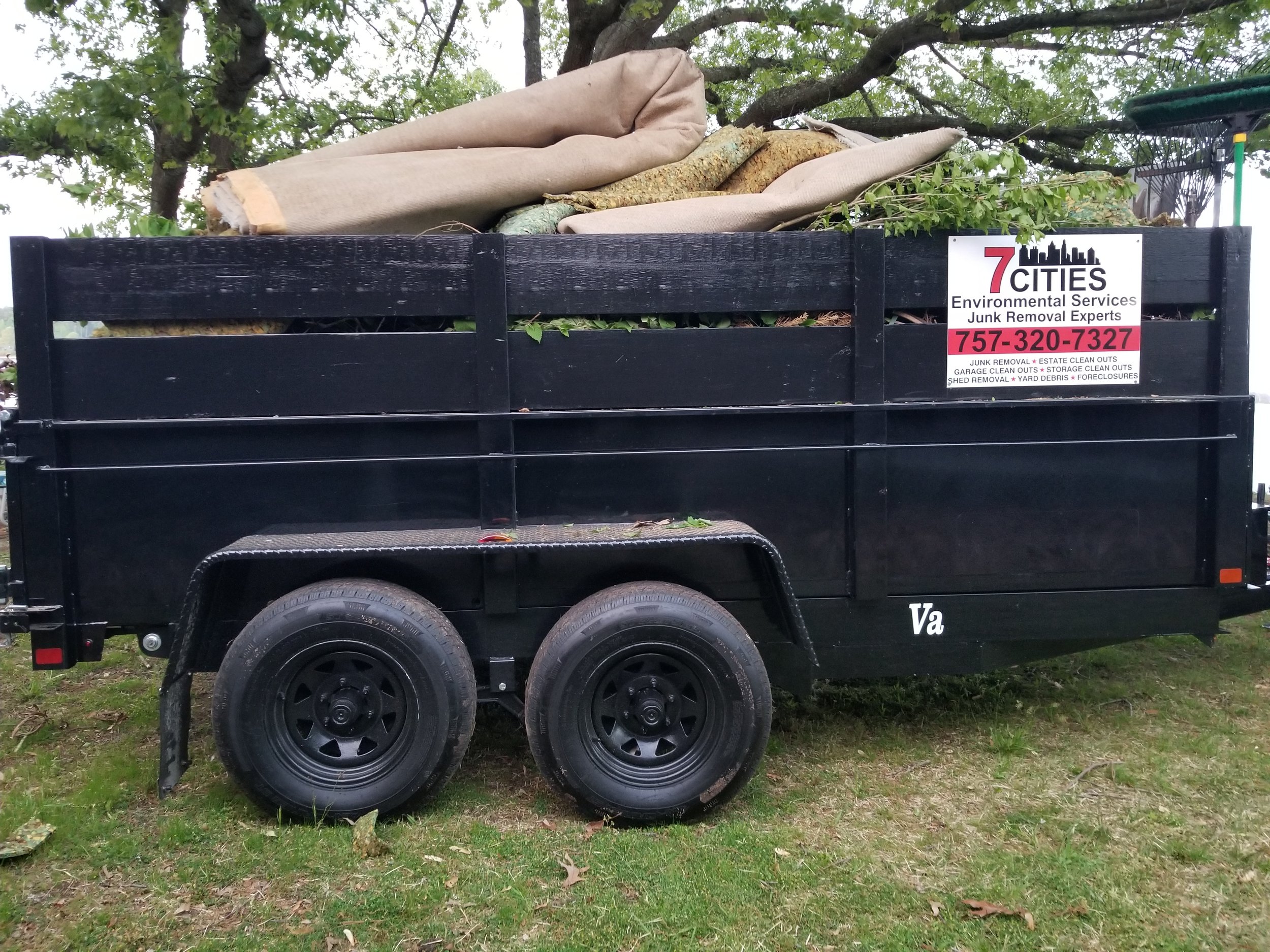 A black trailer filled with yard debris, including tree branches, logs, and rolled-up fabric or carpet, parked on grass under a tree.