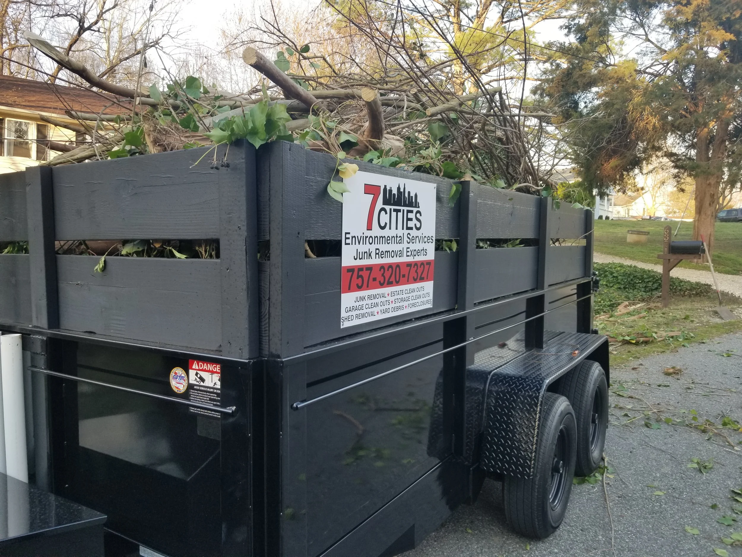 A black utility trailer filled with cut branches and leaves, parked outdoors, with a sign that reads '7 Cities Environmental Services Junk Removal Experts' and a phone number.