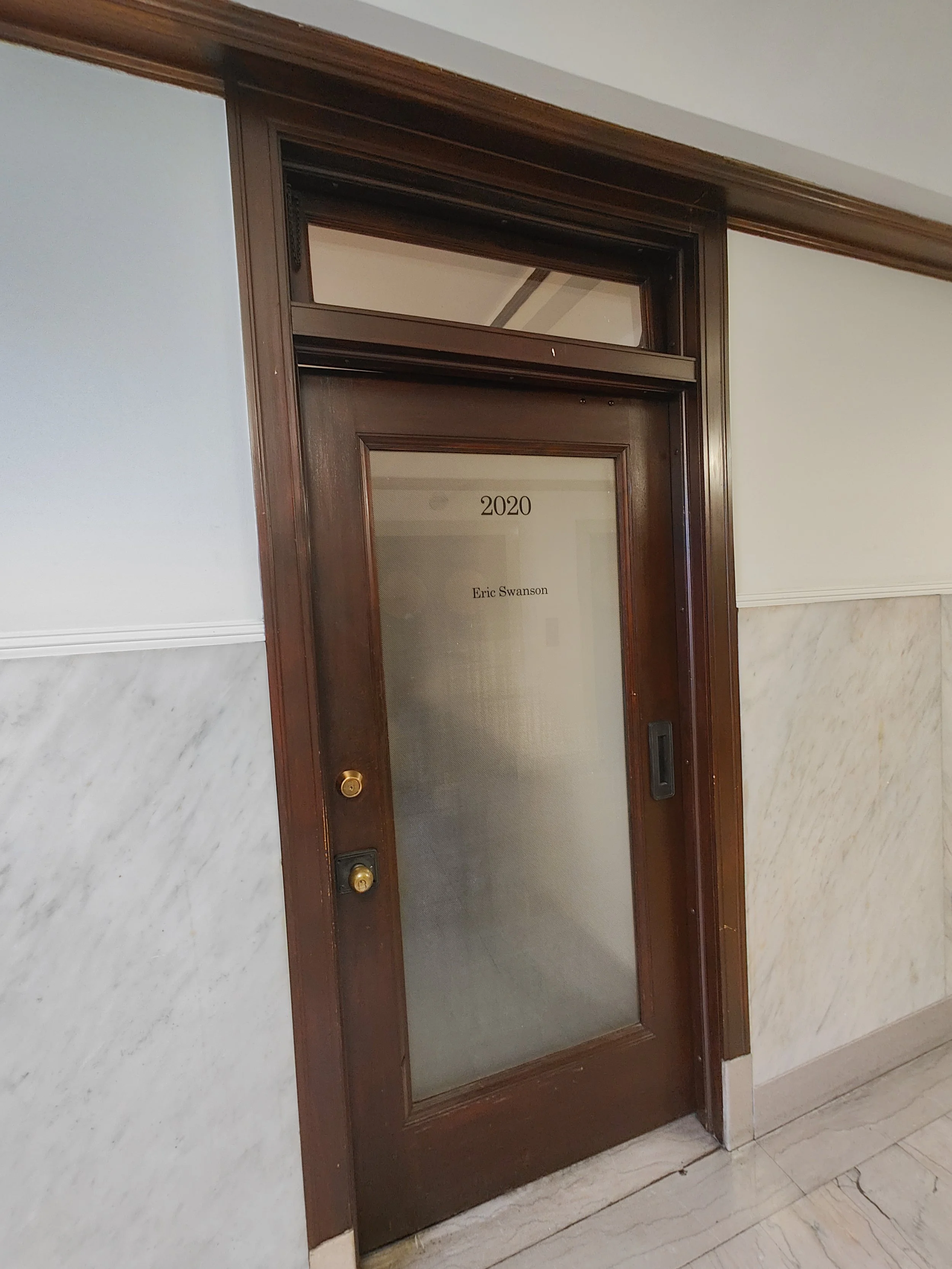 An elevator door with a wooden frame and a frosted glass panel. The panel has the number 2020 and the name Eric Swanson printed on it. The elevator is in a marble-floored hallway with white and beige walls.