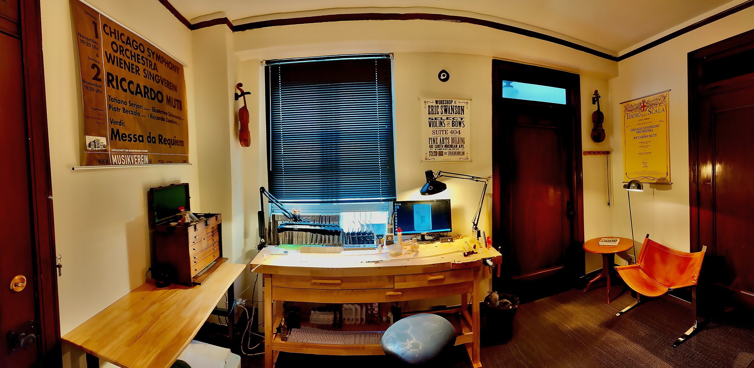 A home office with a wooden desk holding computer monitors, a desk lamp, and various small items. There is a window with blinds behind the desk, two violins hanging on the wall, and framed posters on the walls. One poster lists concert details, and another advertises a workshop. A small orange chair and a round table are also in the room.