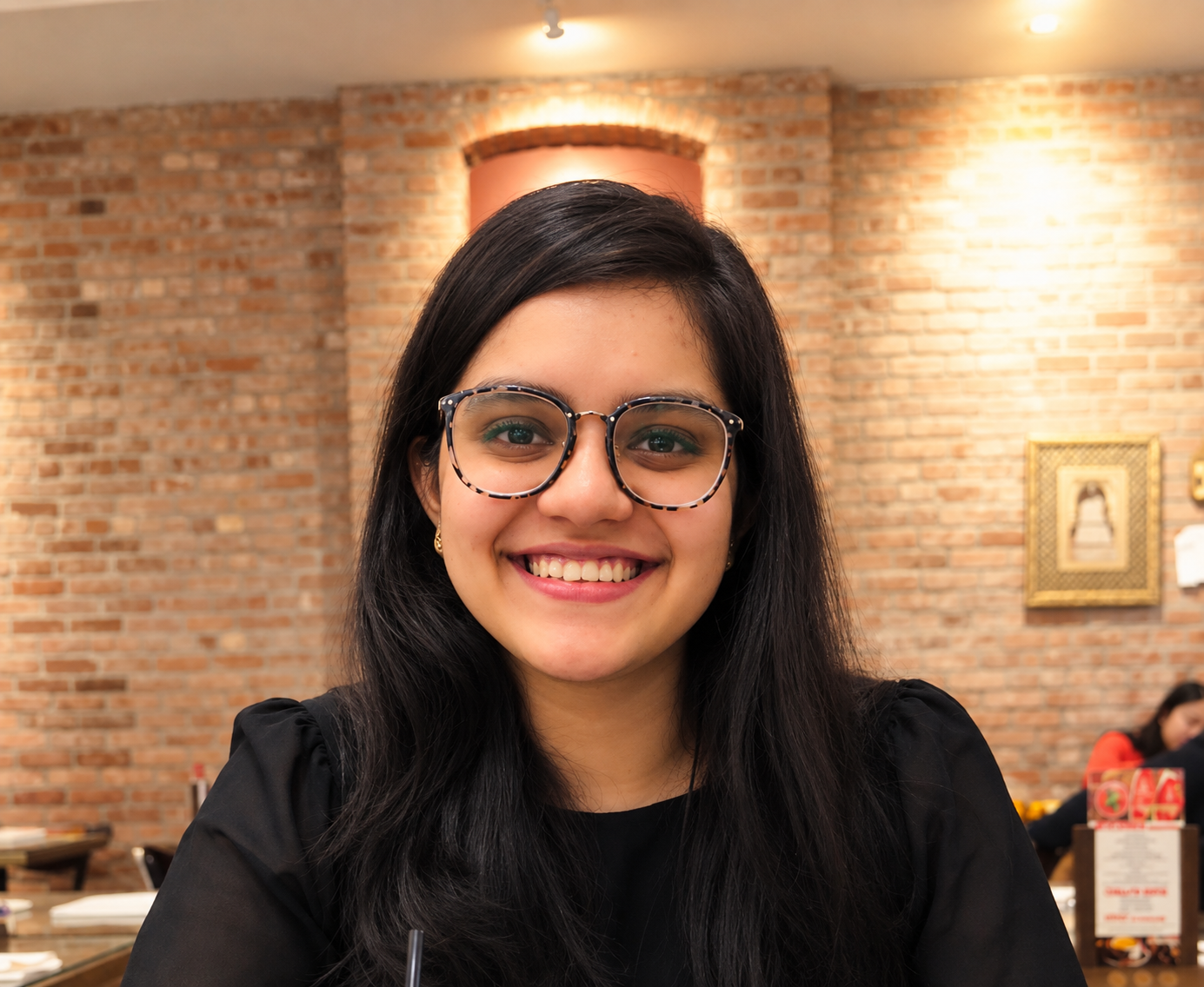 A smiling woman with long black hair, wearing eyeglasses and a black top, sitting in a restaurant with a brick wall background.