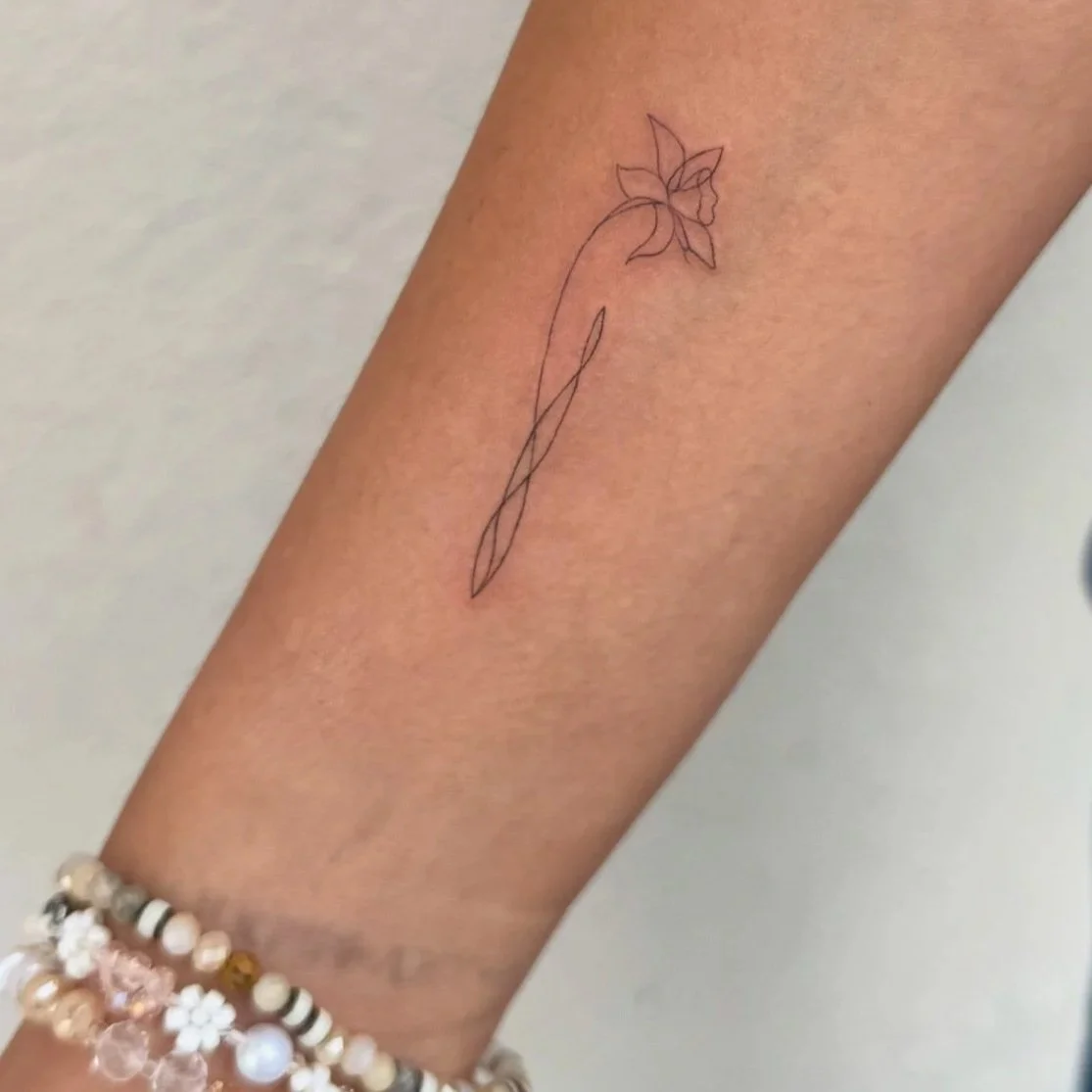 Minimalist fine‑line flower tattoo by Madison Davenport featuring a single delicate stem with pointed petals, drawn in clean black ink; elegant, modern botanical design emphasizing simplicity and subtle detail.