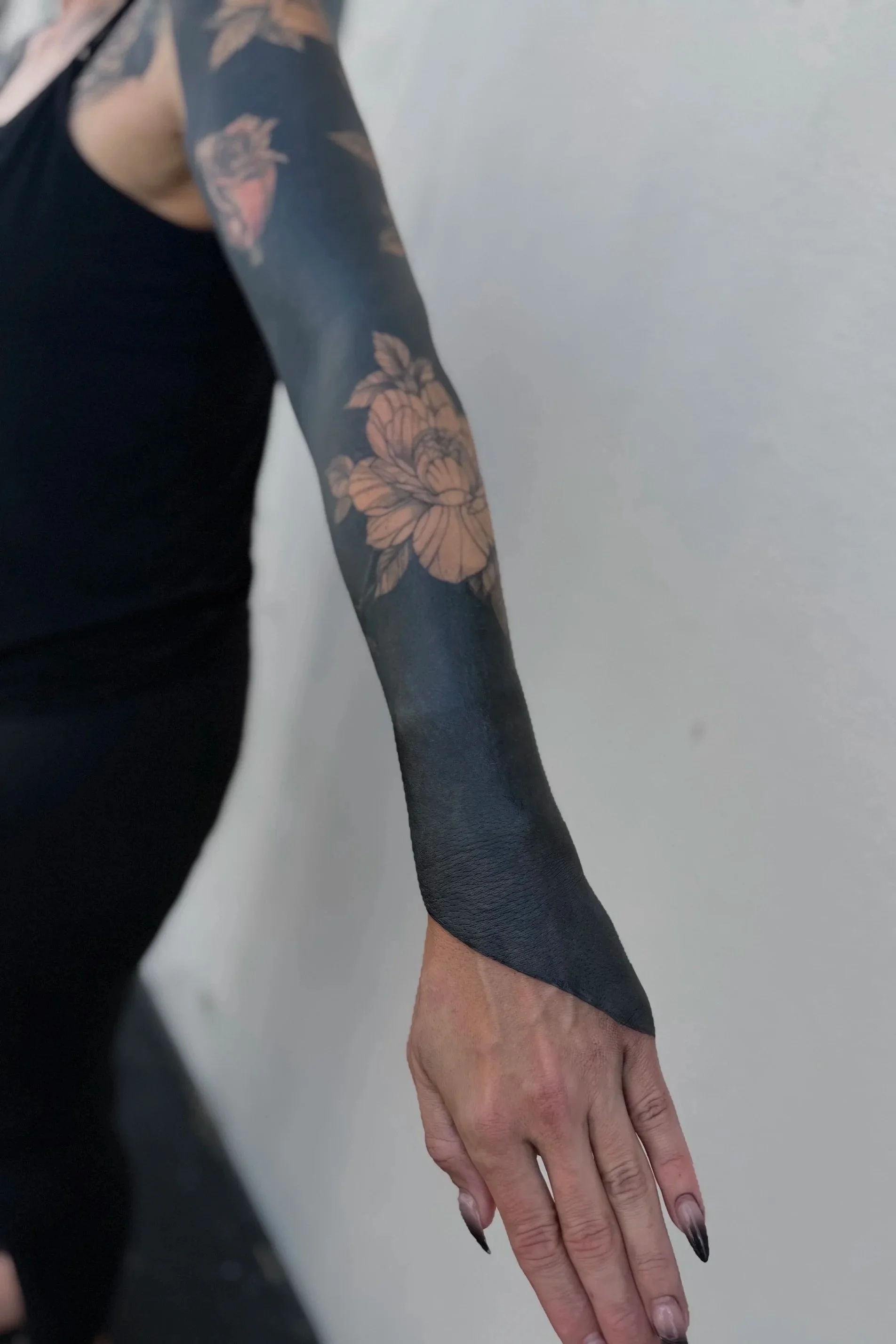 Blackout floral sleeve tattoo by Kelly Cunningham featuring large floral blooms left in negative space against a solid blackwork background extending from the forearm to the hand.