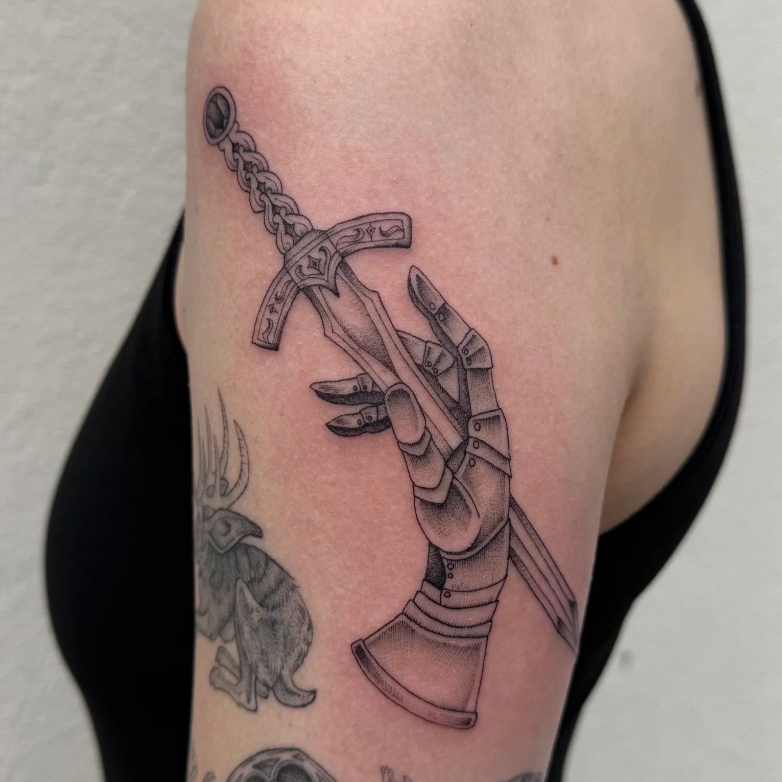 Black-and-grey tattoo by Madison Davenport at Handsome Devil Tattoo in Saddle Brook NJ featuring an armored hand gripping a detailed sword hilt with intricate metallic shading; high-contrast illustrative style with fine detail and texture.