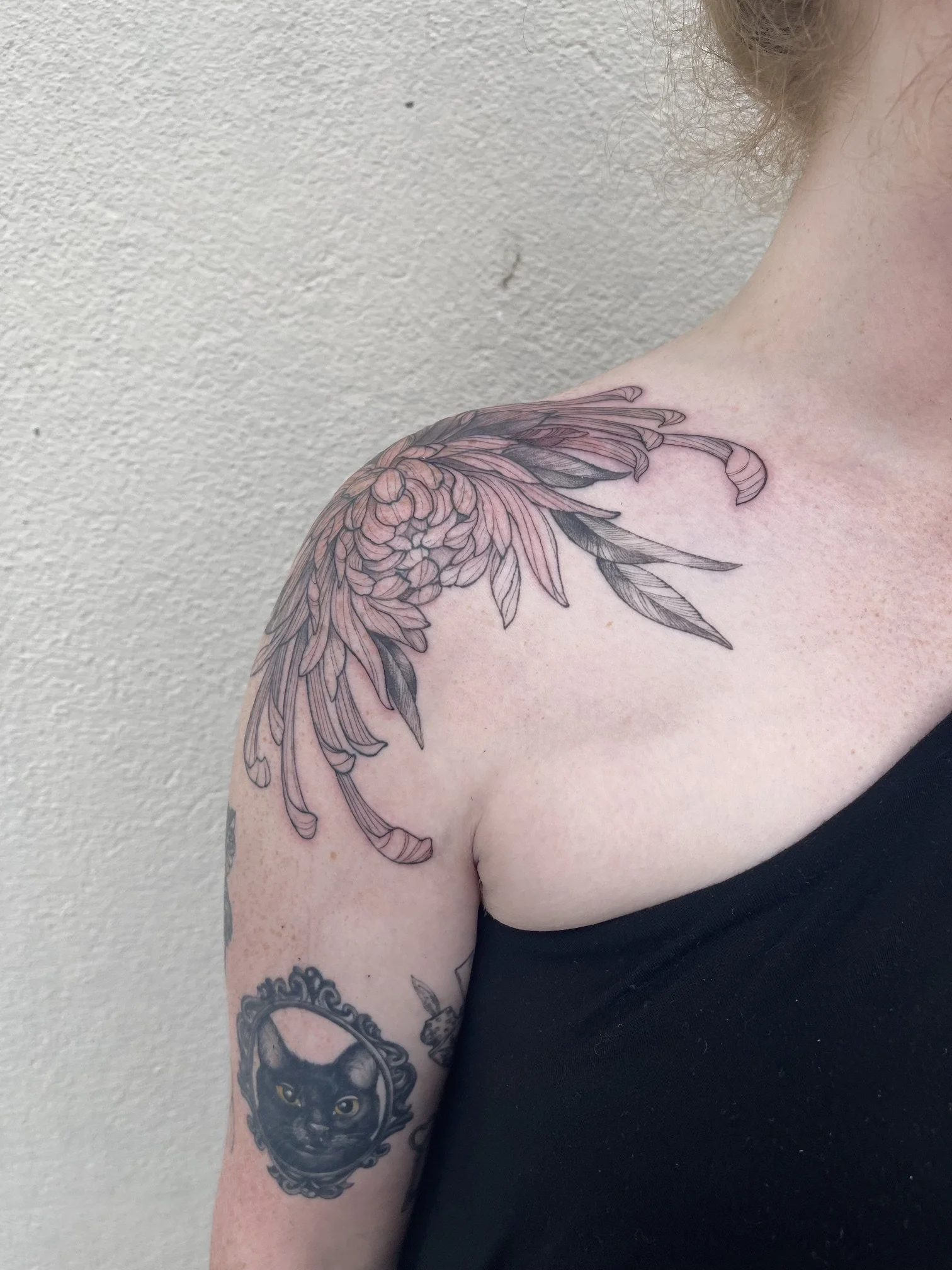 Close-up of a person's shoulder and upper arm with a floral tattoo featuring pink and black ink. Part of a black lion face tattoo is visible on the lower arm. The person is wearing a black sleeveless top, and the background is a plain beige wall.