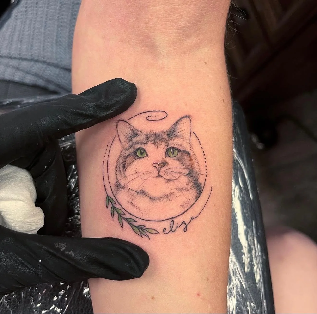 Tattoo of a cat's face with green eyes, decorative lines, a branch with leaves, and the name Elise written in cursive.