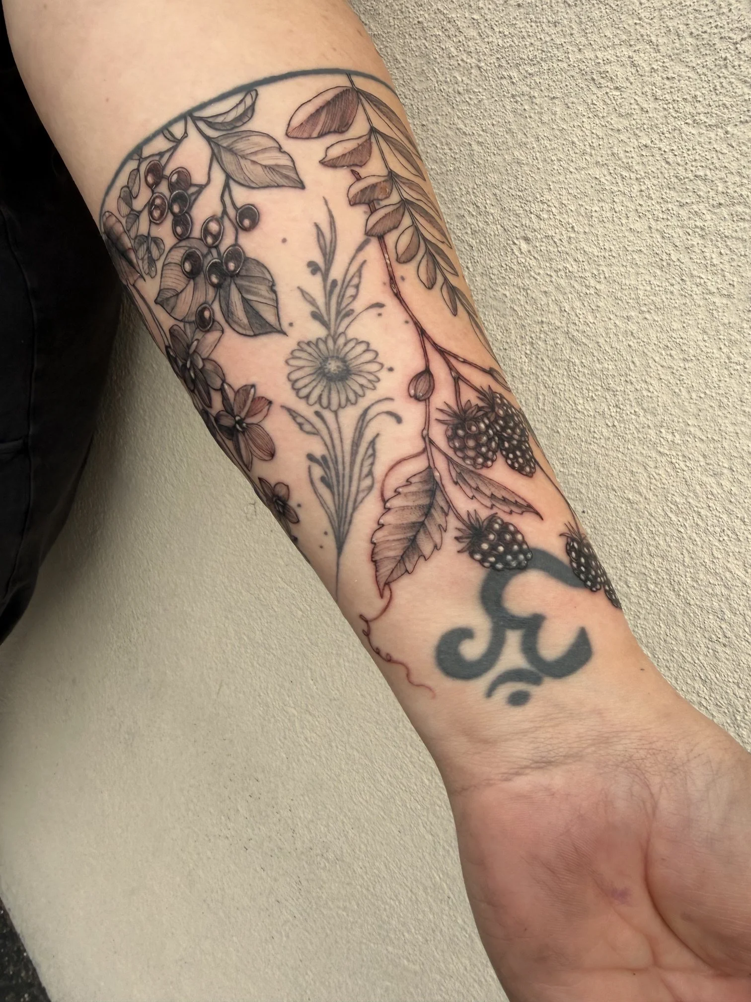 A tattoo on a person's forearm featuring various plants and berries, with black outlines and some shading, and a word or name partially visible below.
