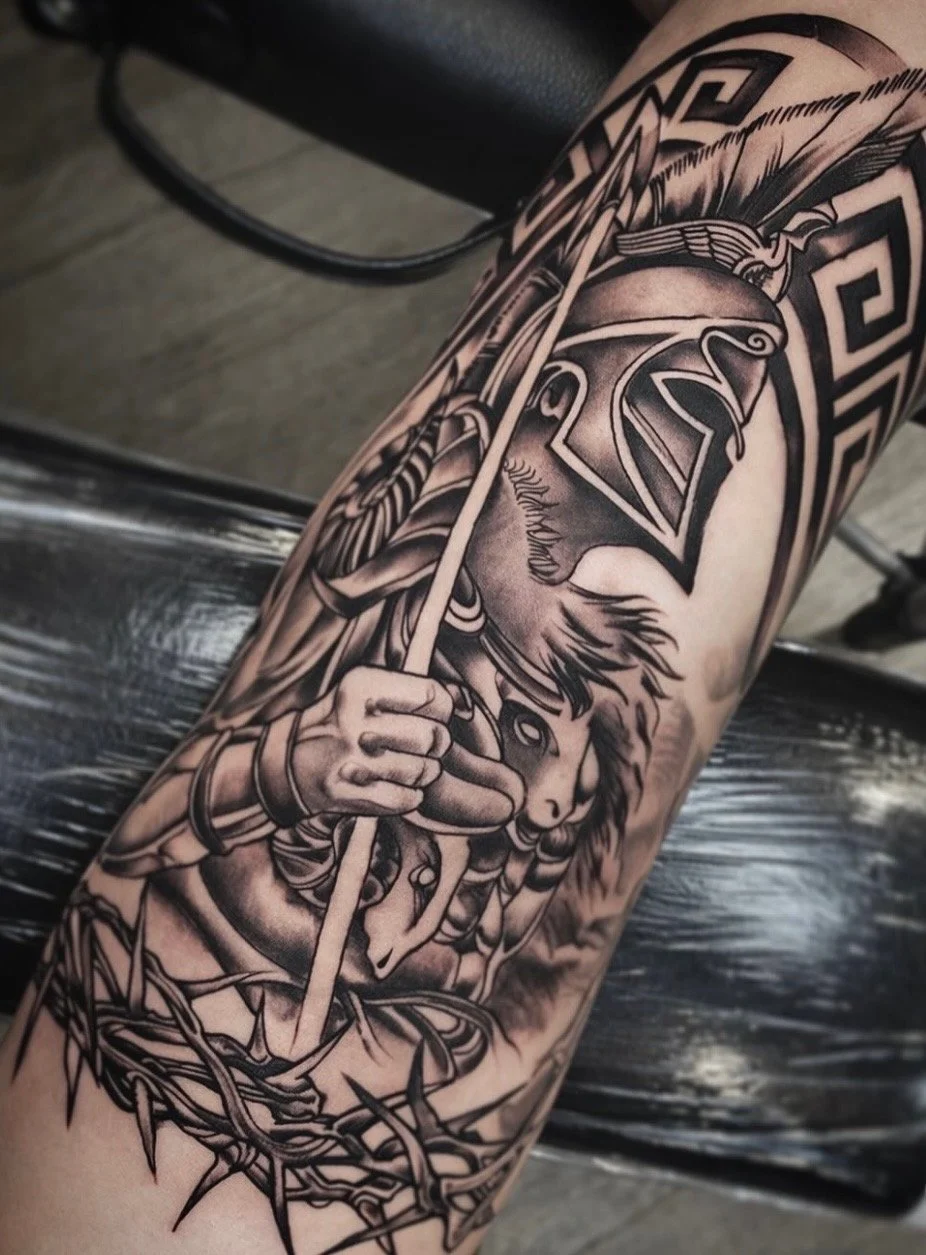 Black and gray tattoo of a warrior princess with a helmet, holding a spear and a shield, on a person's arm.