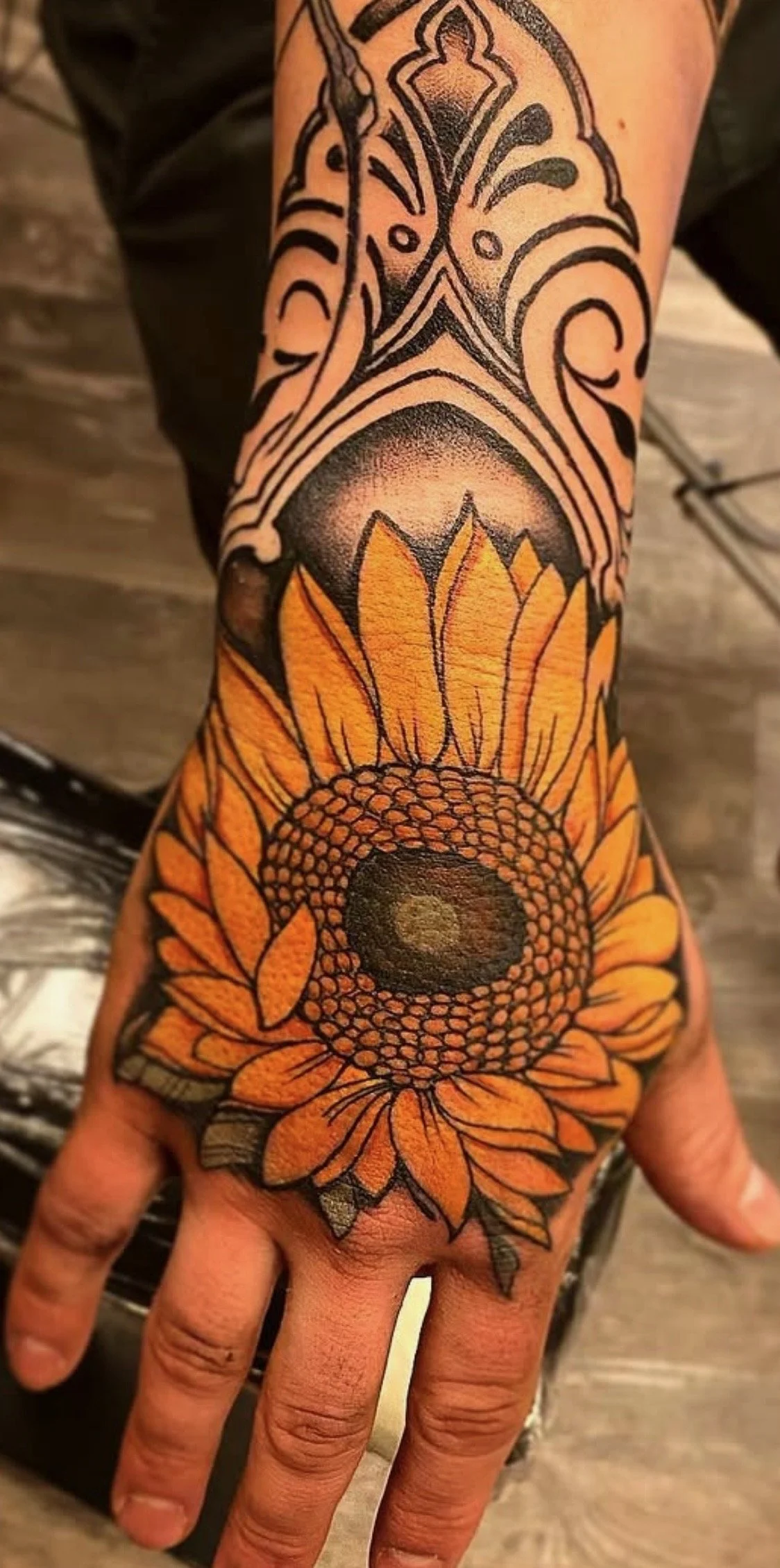 Tattoo of a sunflower on a person's hand and forearm featuring orange petals, black outlines, and detailed shading.