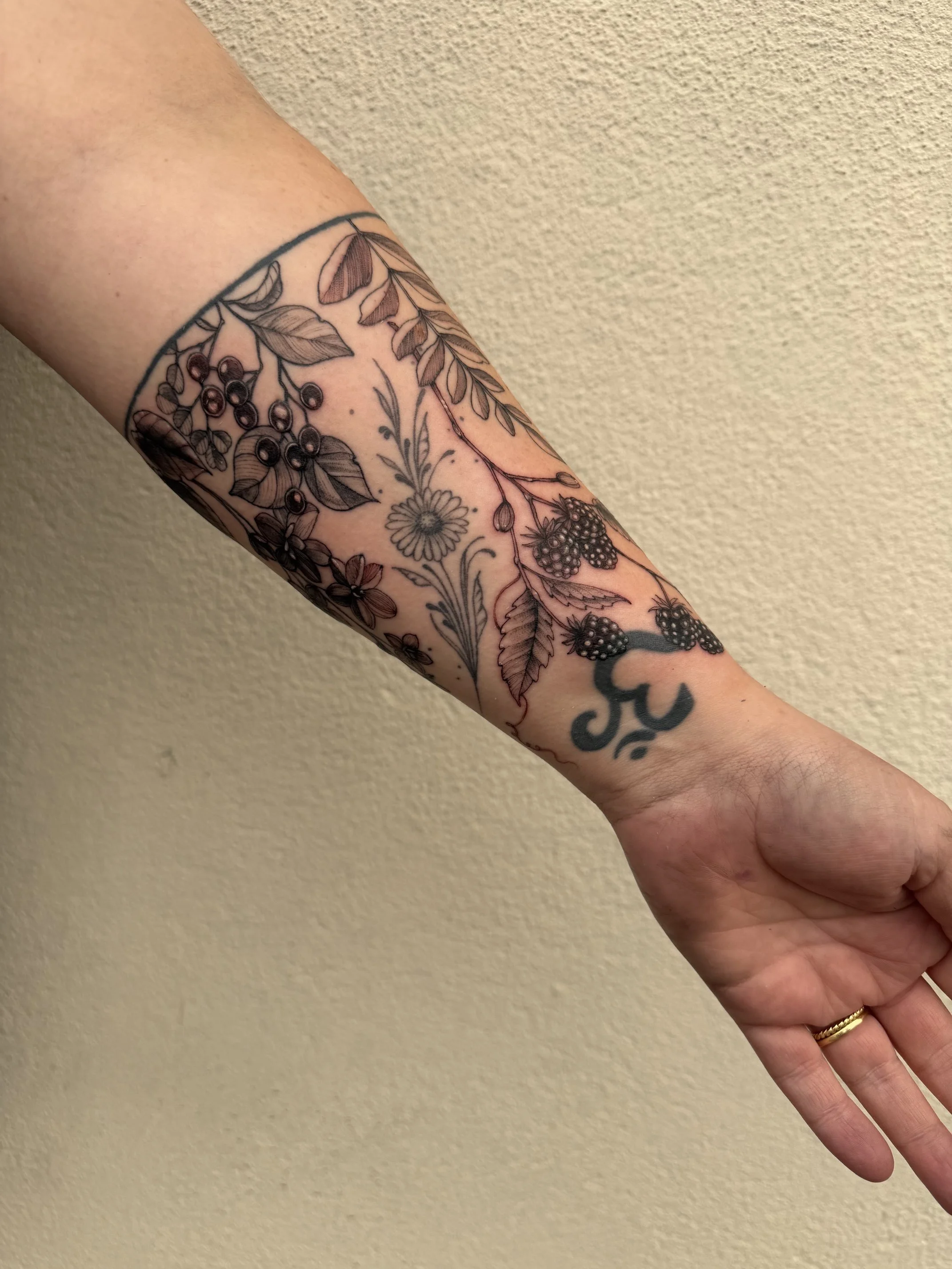 A tattooed arm with floral and berry designs, including leaves, berries, and a flower, against a beige wall.