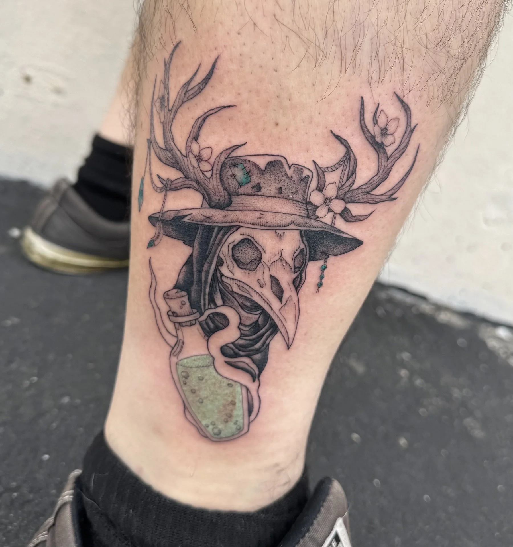 Black and grey plague doctor tattoo featuring a bird-beaked mask, wide-brimmed hat, antlers with blossoms, and a potion bottle with subtle color accents.