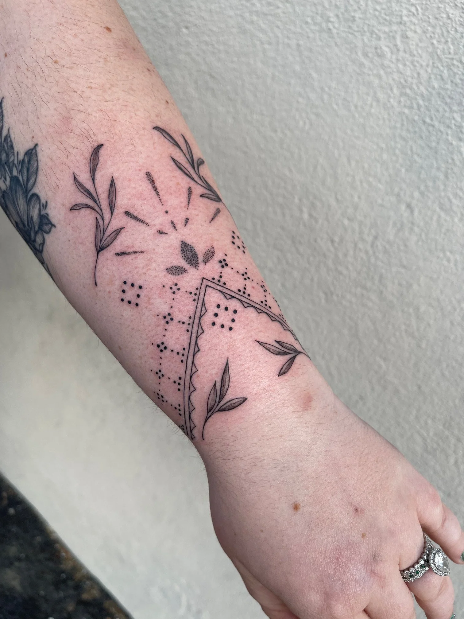 A person's forearm with a black ink tattoo of leaves, dotted patterns, and geometric shapes, with rings on the fingers.