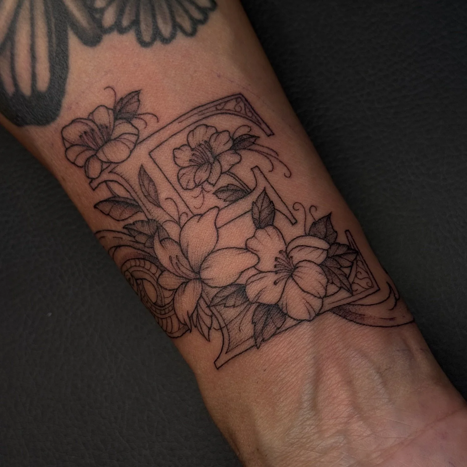 Line drawing tattoo of flowers and a decorative border on a person's forearm.