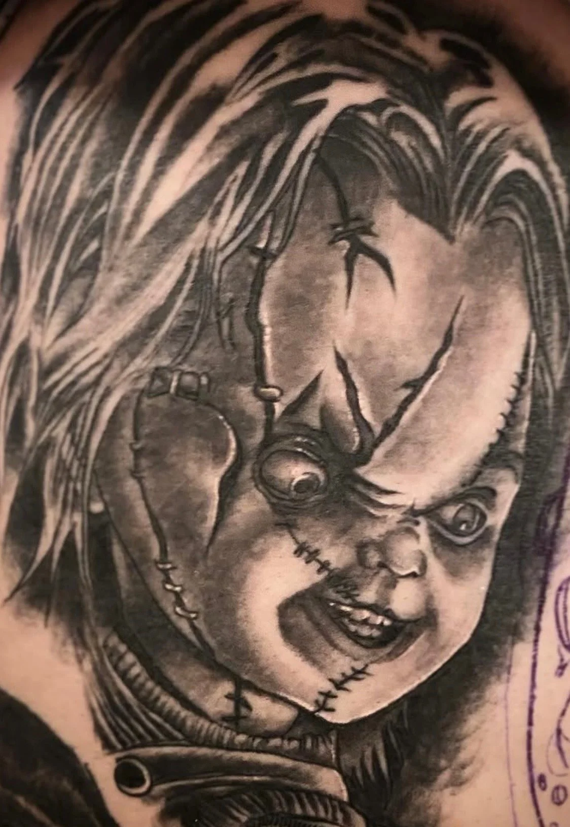 A tattoo of a woman with long hair, with her eyes closed, and a sinister clown face with a stitched mouth and dark eyes, integrated into her face.