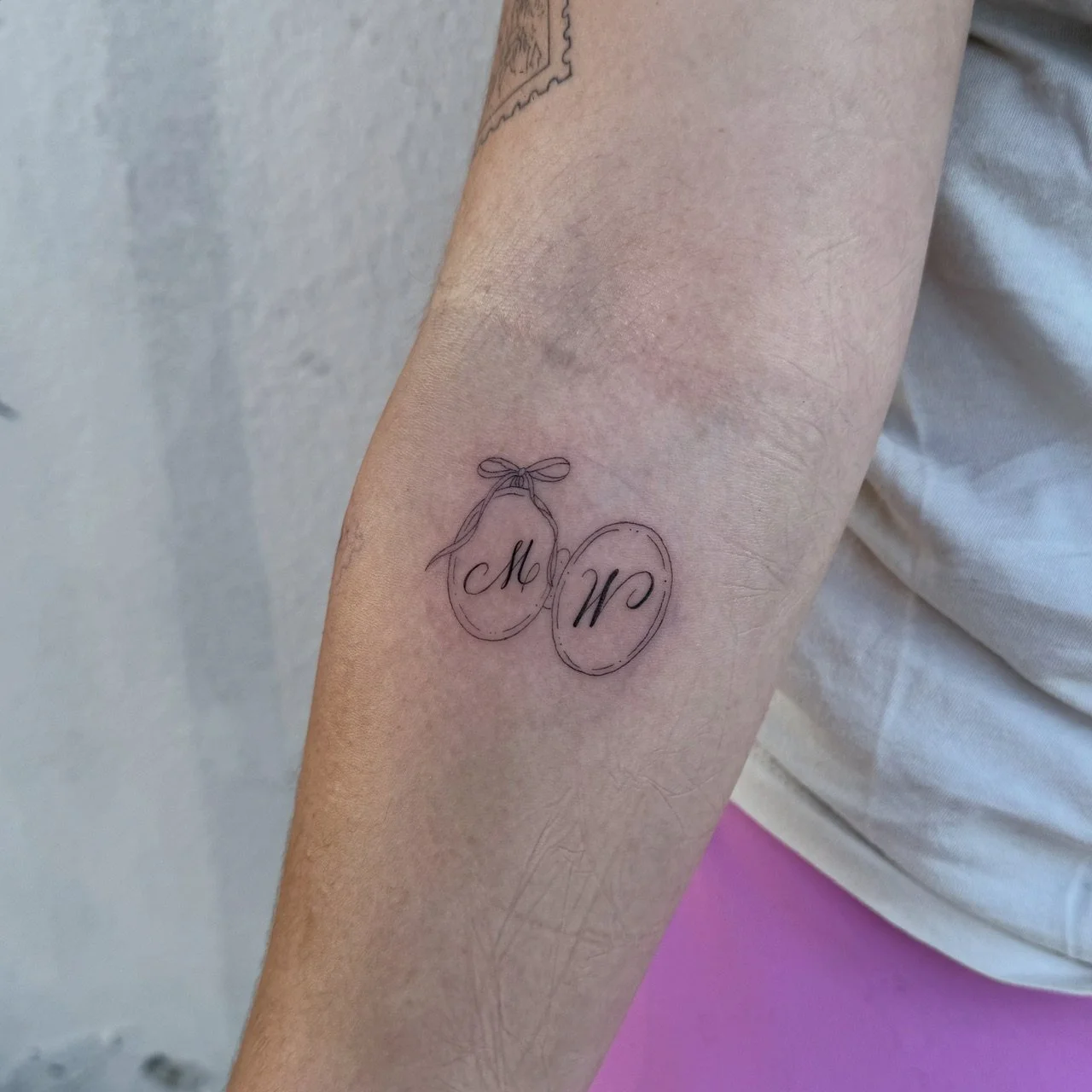 Fine line initial tattoo by Madison Davenport featuring the letters M and W inside decorative egg-shaped frames connected by a delicate bow.
