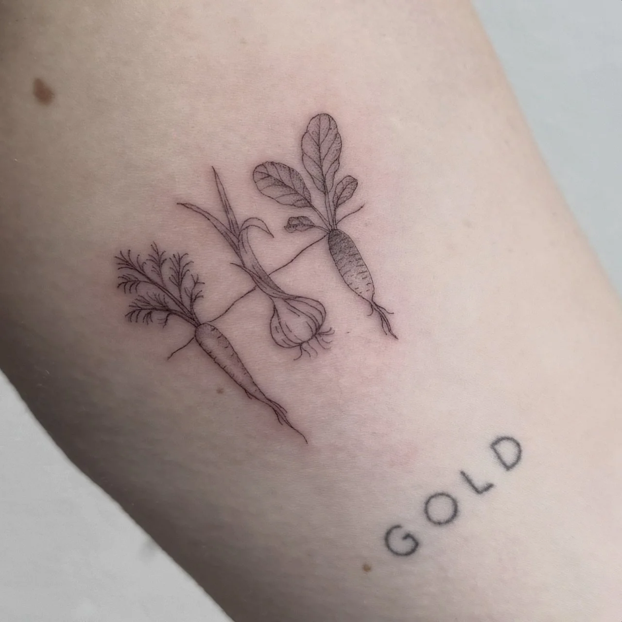 A minimalist black ink tattoo of two carrots with leaves, arranged diagonally, with the word 'GOLD' written below.