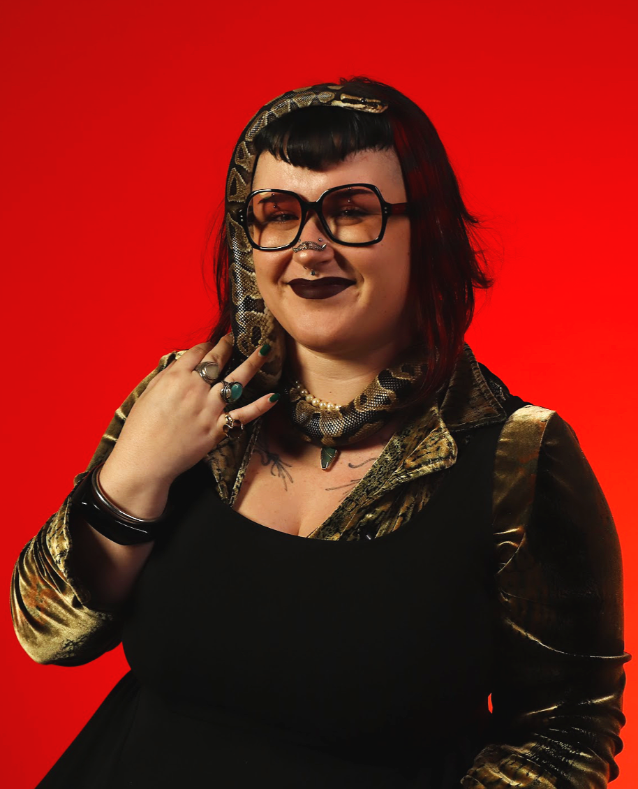 Headshot of Tuesday with dark hair, black glasses, dark lipstick, and facial piercings, poses with a snake on her neck and shoulder against a red background dressed in a black top and metallic gold jacket, with rings, bracelets, and a necklace.