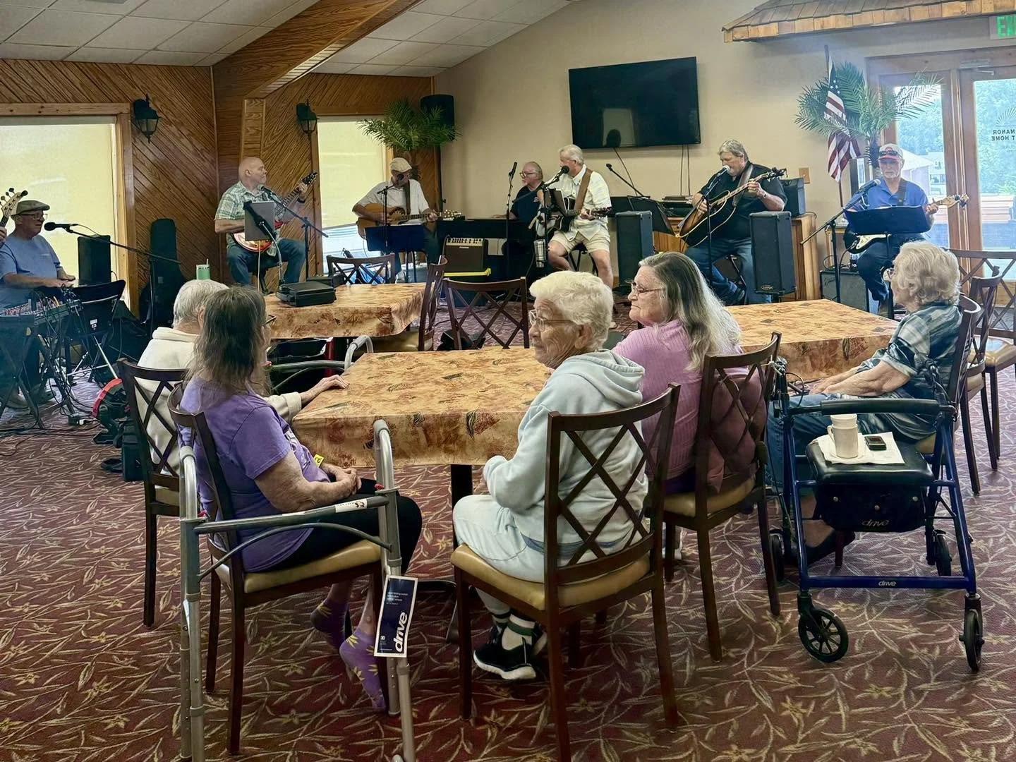 A musical performance by a band with guitars and keyboard on a stage in a restaurant or community center, with elderly audience members sitting at tables, some using walkers or wheelchairs, watching the performance.