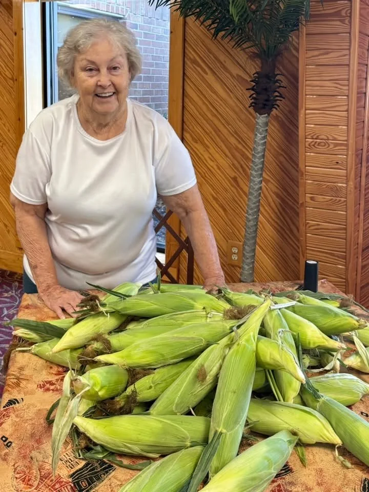 An elderly woman standing behind a table with a large pile of fresh ears of corn in husks, inside a room with wooden paneling and a large potted plant nearby.