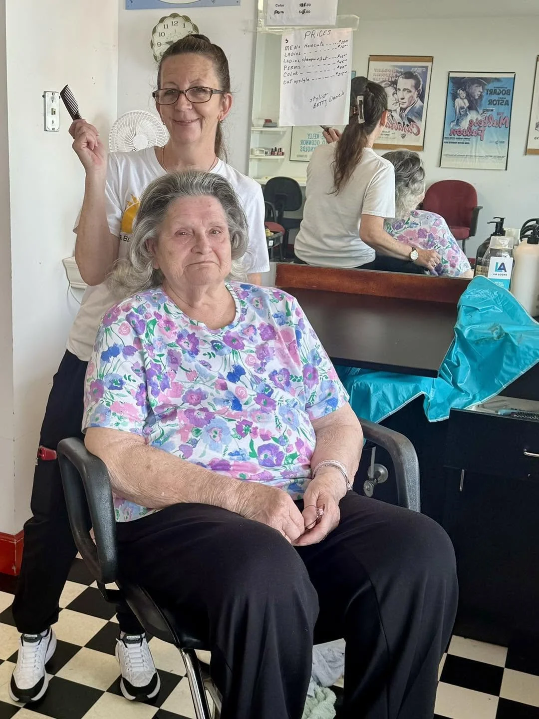 A hairstylist standing behind an elderly woman sitting in a black salon chair in a hair salon. The elderly woman has gray hair, wearing a floral shirt, and has a serious expression. The hairstylist has glasses, is holding a black comb, and is smiling