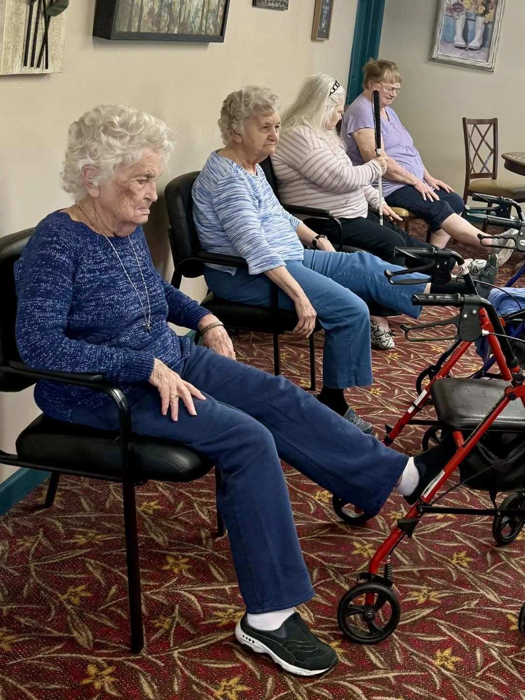Four elderly women sitting in chairs, in a room with framed artwork and patterned carpet.