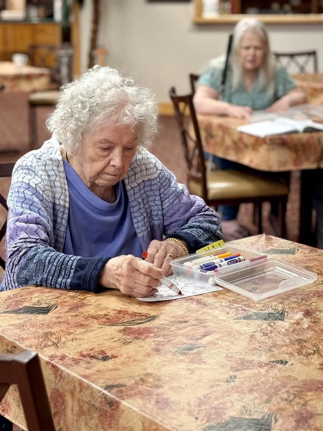 An elderly woman sitting at a table coloring with markers in a care home or community center with another woman sitting at a nearby table in the background.