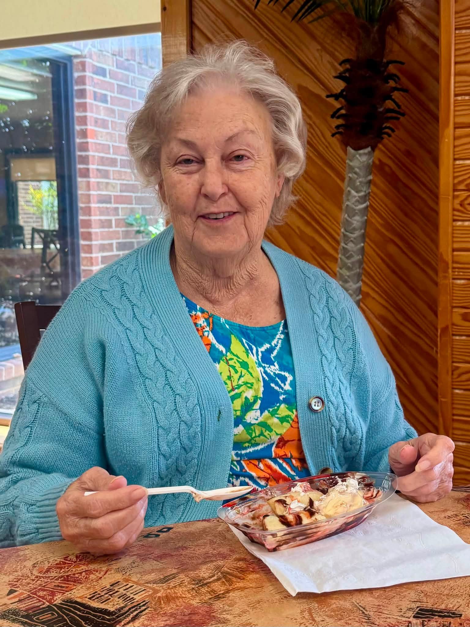 An elderly woman with gray hair, wearing a blue sweater and colorful dress, is sitting at a wooden table holding a spoon and a bowl filled with ice cream or dessert. She is smiling and appears to be enjoying her treat inside a cozy room with wooden p